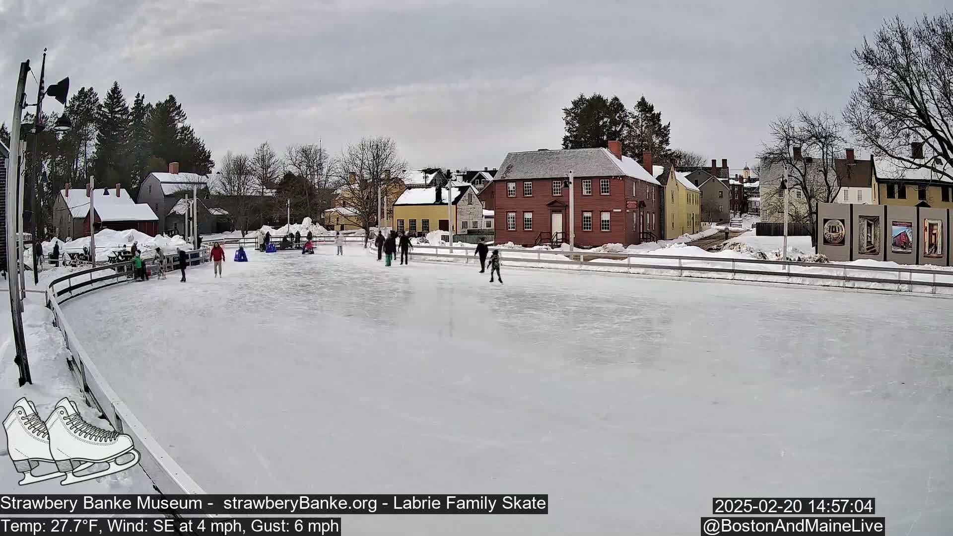 A mostly empty outdoor ice skating rink surrounded by snow-covered historic buildings on an overcast day with a southeast wind.
