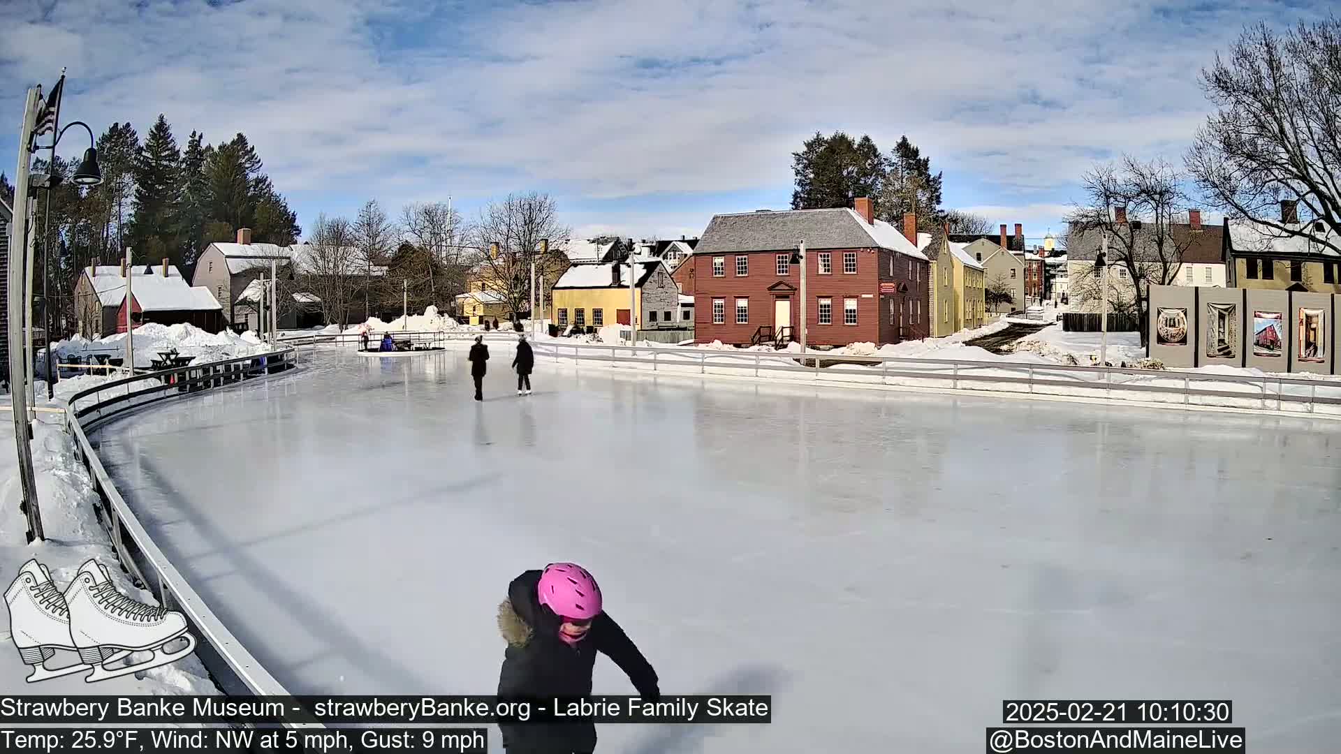 A partly sunny day with a northwest wind shows several people ice skating on an outdoor rink in front of a row of historic buildings, which are covered in snow.