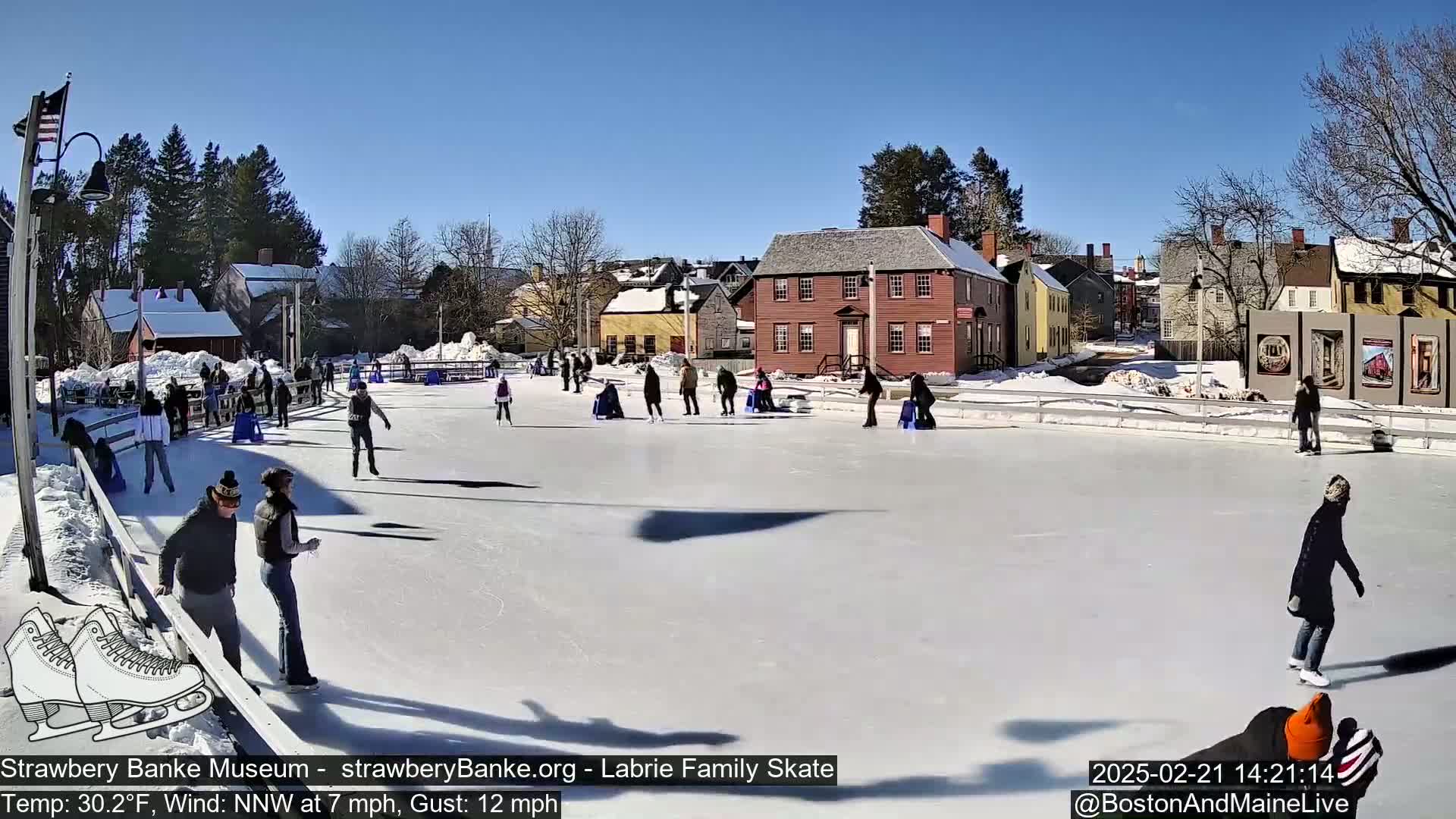 People ice skate on an outdoor rink on a sunny day with a light wind.