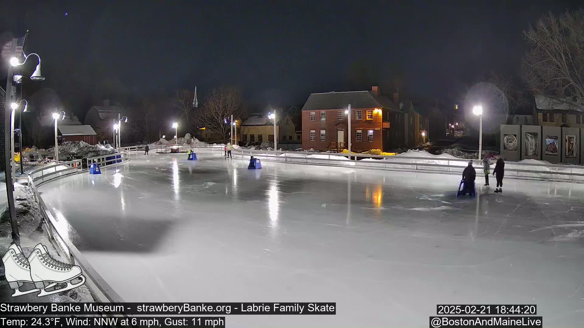 An outdoor ice skating rink at night, illuminated by lights, with several people ice skating and others using sleds, under a clear, dark sky.