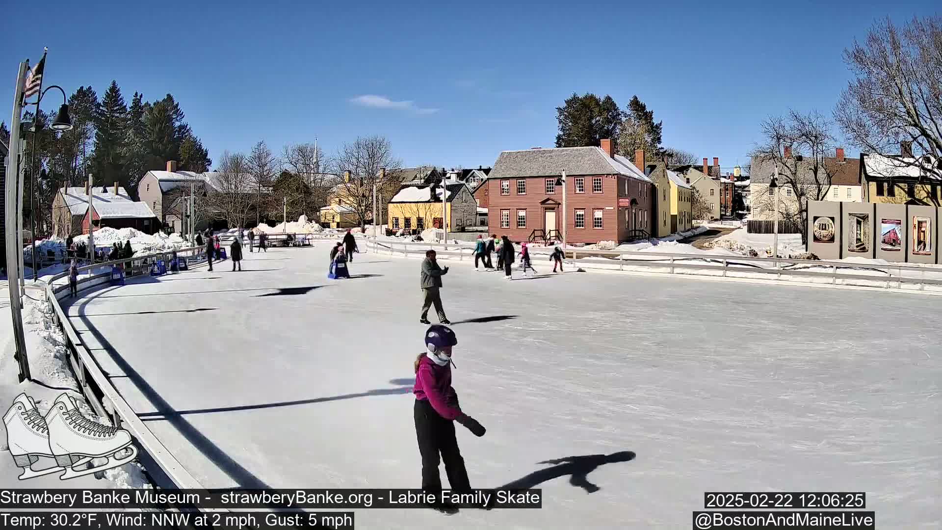 People ice skate on an outdoor rink in front of historic buildings on a sunny day with a light wind.