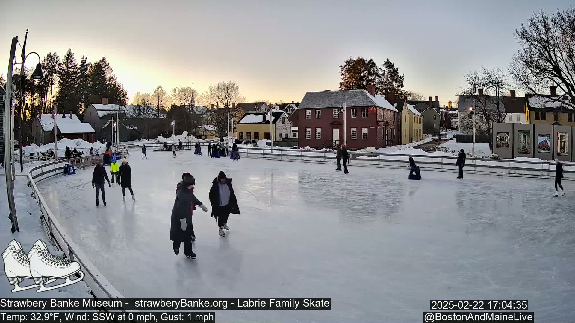People ice skate on an outdoor rink at sunset, surrounded by historic buildings and snow-covered ground.
