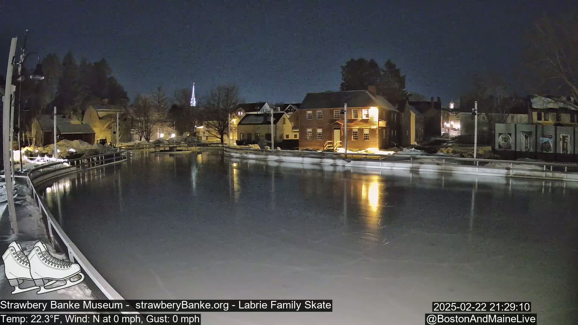 A nighttime view of an outdoor ice skating rink, illuminated by nearby buildings under a clear, dark sky.