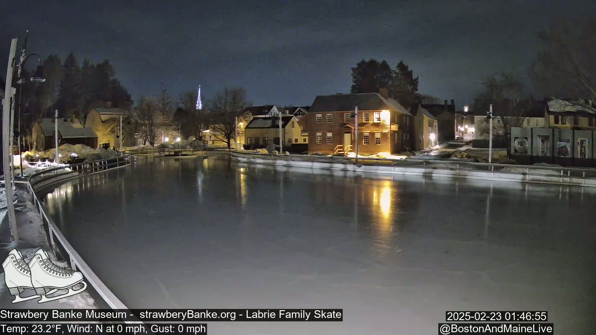 A nighttime view of an outdoor ice skating rink, illuminated by lights, in front of a row of historic-looking buildings under a clear, dark sky.