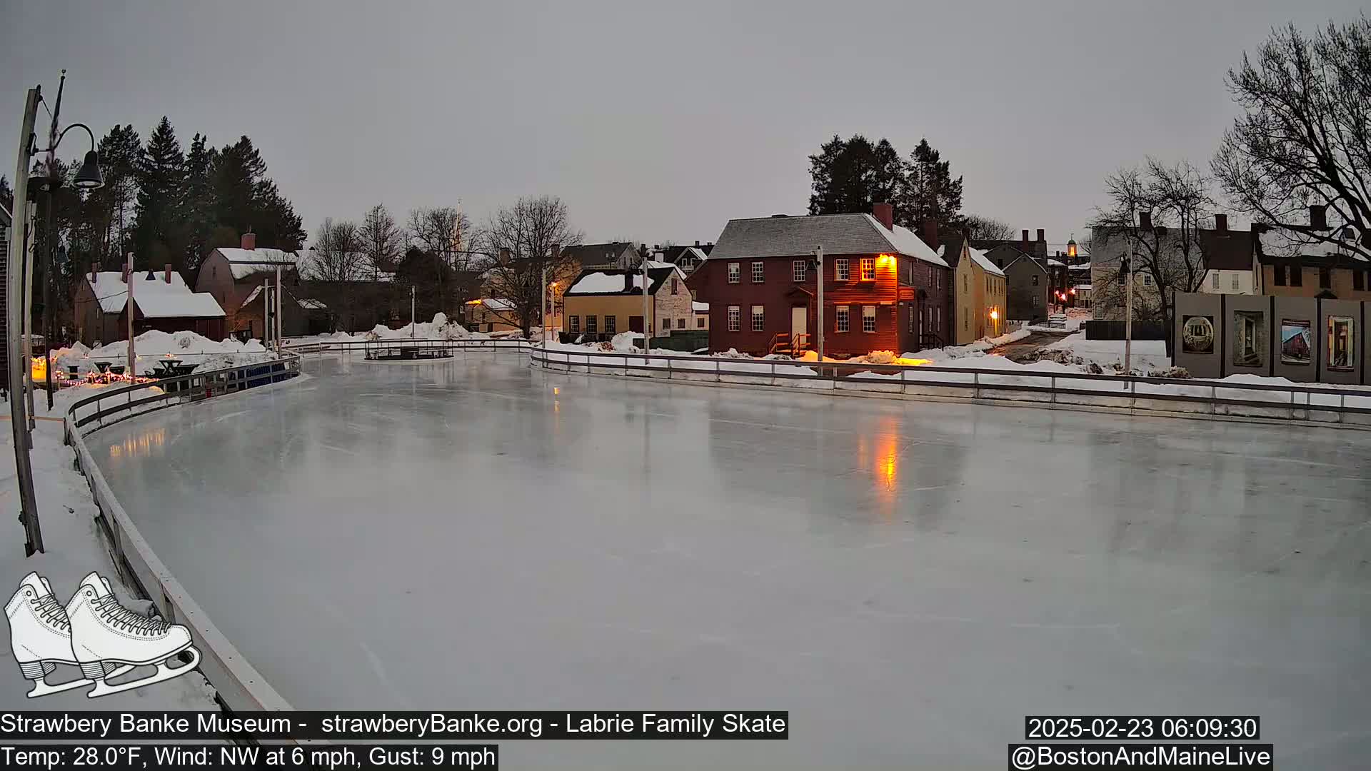 An outdoor ice skating rink, surrounded by snow-covered buildings and trees under a twilight sky, with a northwest wind.