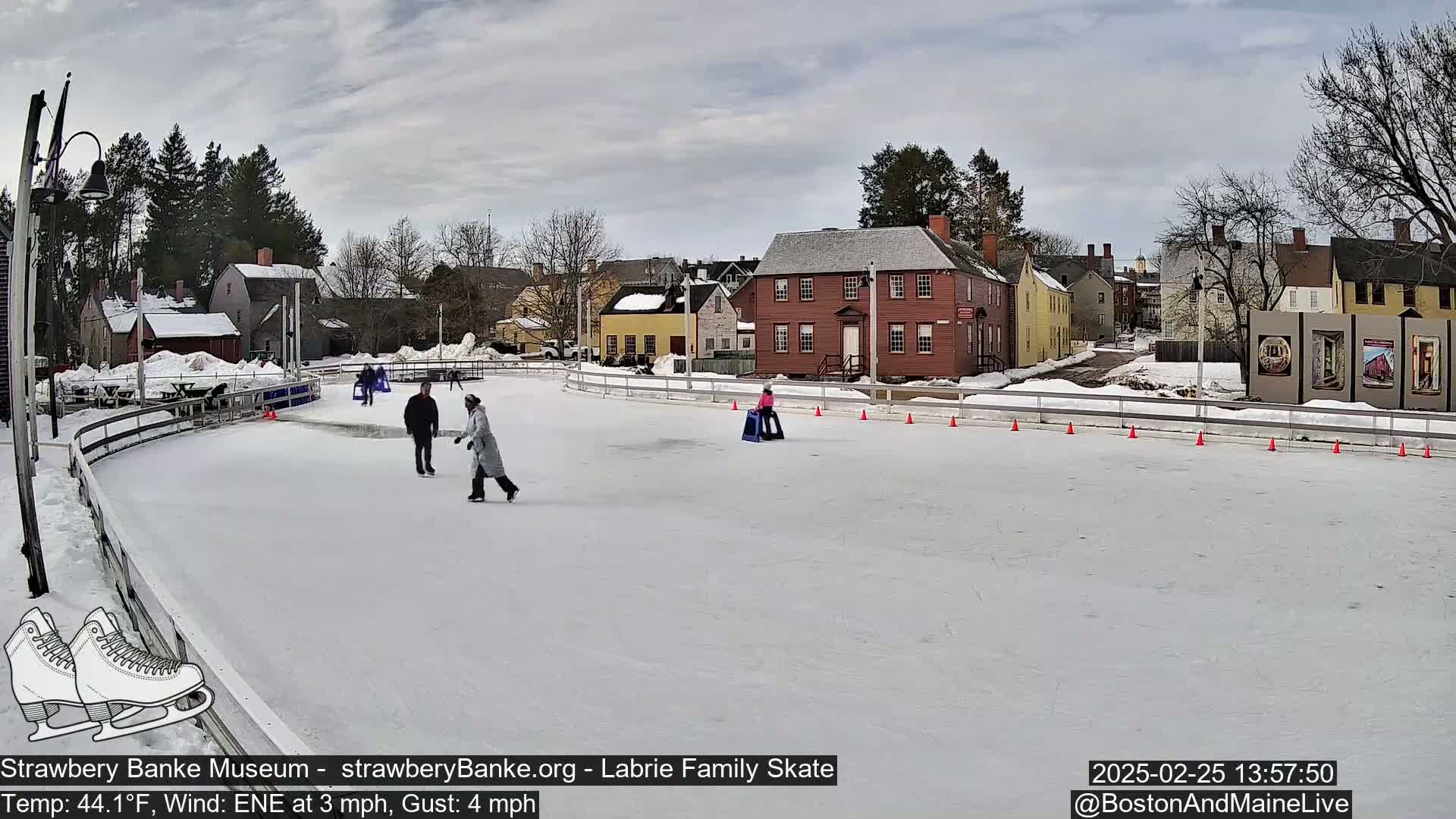 A snow-covered outdoor ice rink with several people ice skating, surrounded by historic buildings under a cloudy sky.