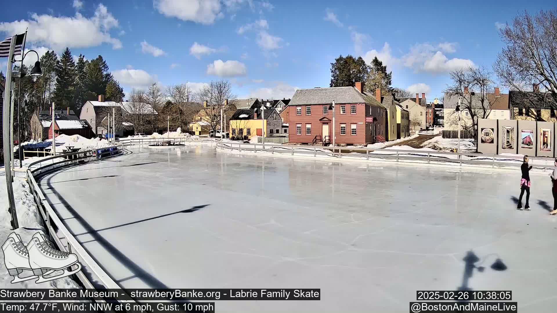 A mostly sunny day shows an outdoor ice skating rink surrounded by historic buildings, with two people ice skating.