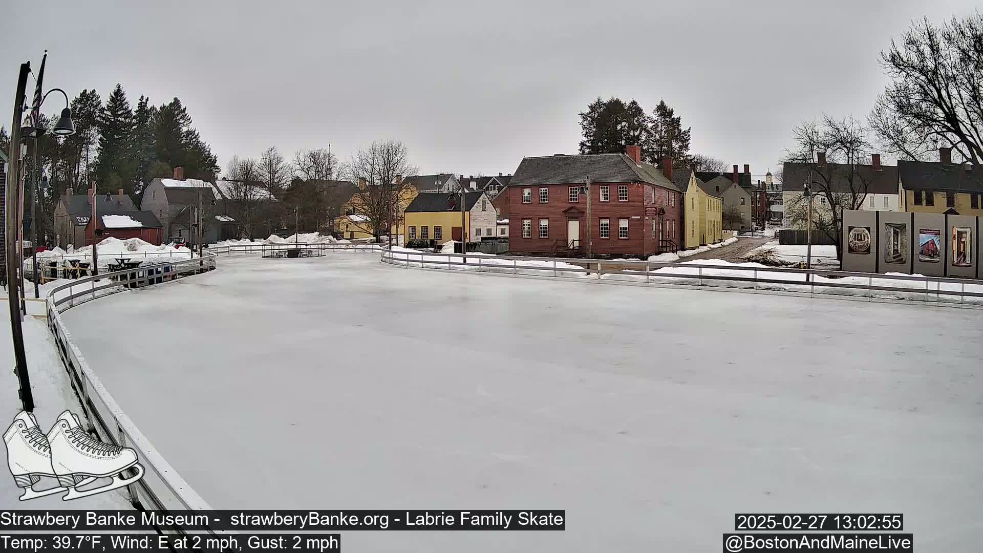 A snow-covered outdoor ice rink is situated in front of several historic-looking buildings on an overcast day with a light east wind.