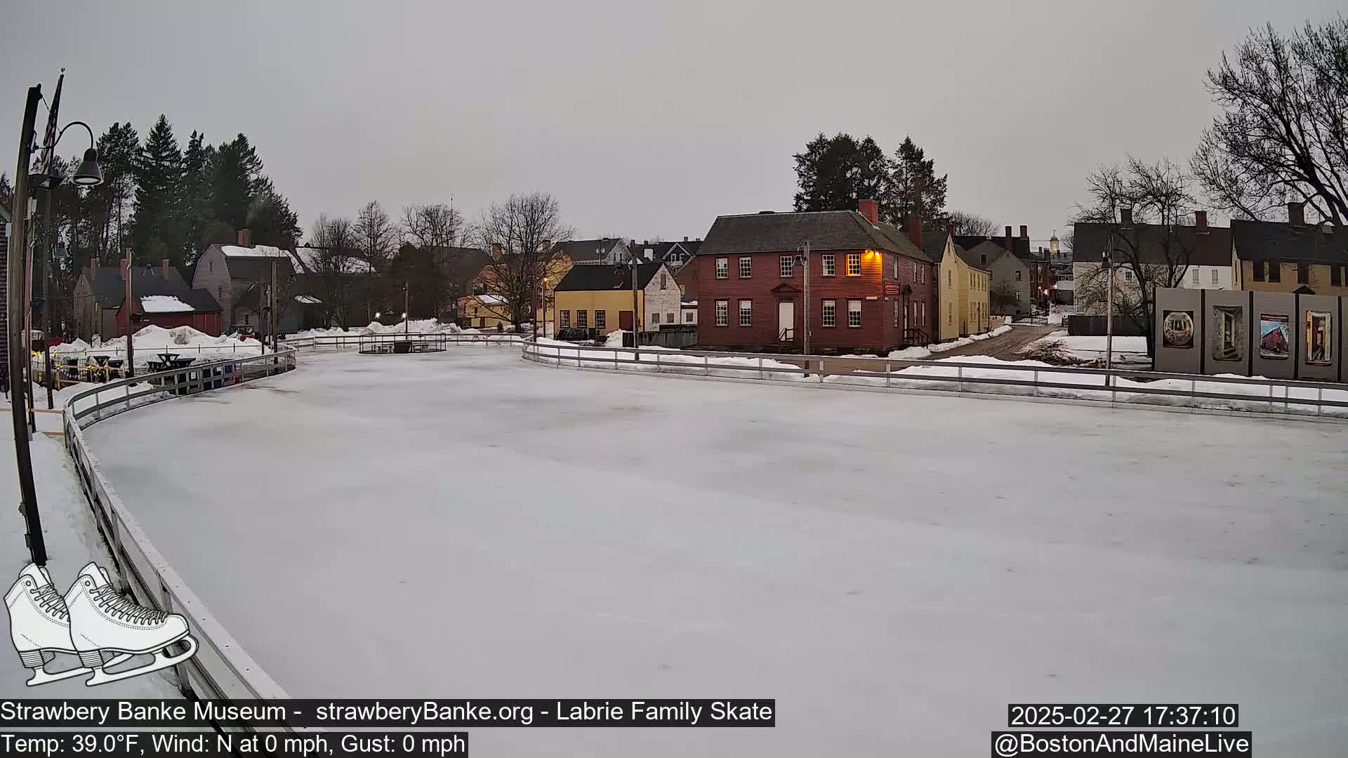 A snow-covered outdoor ice rink sits before a row of historic buildings under an overcast sky.