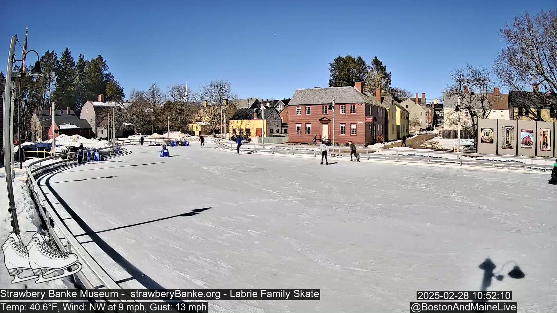 A sunny day shows several people ice skating on an outdoor rink in front of historical-looking buildings.