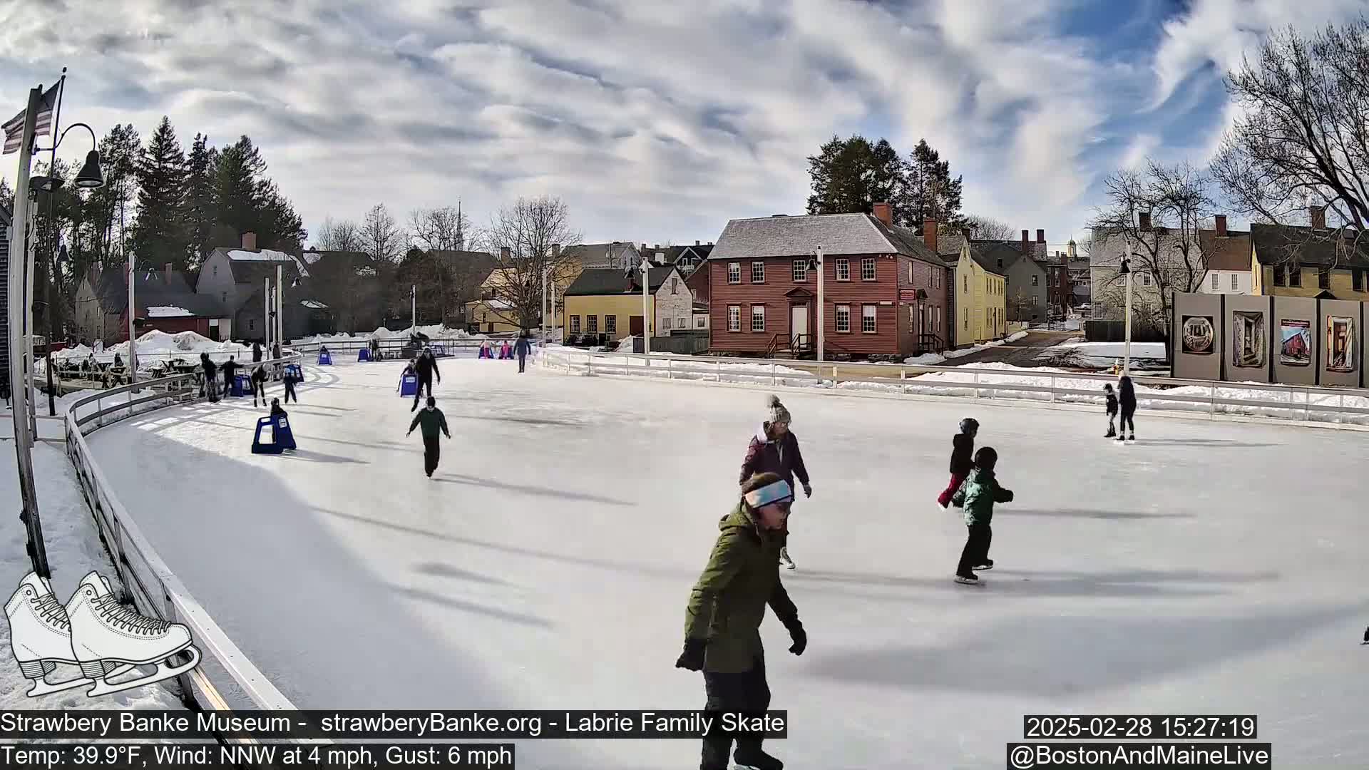 People ice skate on an outdoor rink under a partly cloudy sky, with buildings and trees in the background.