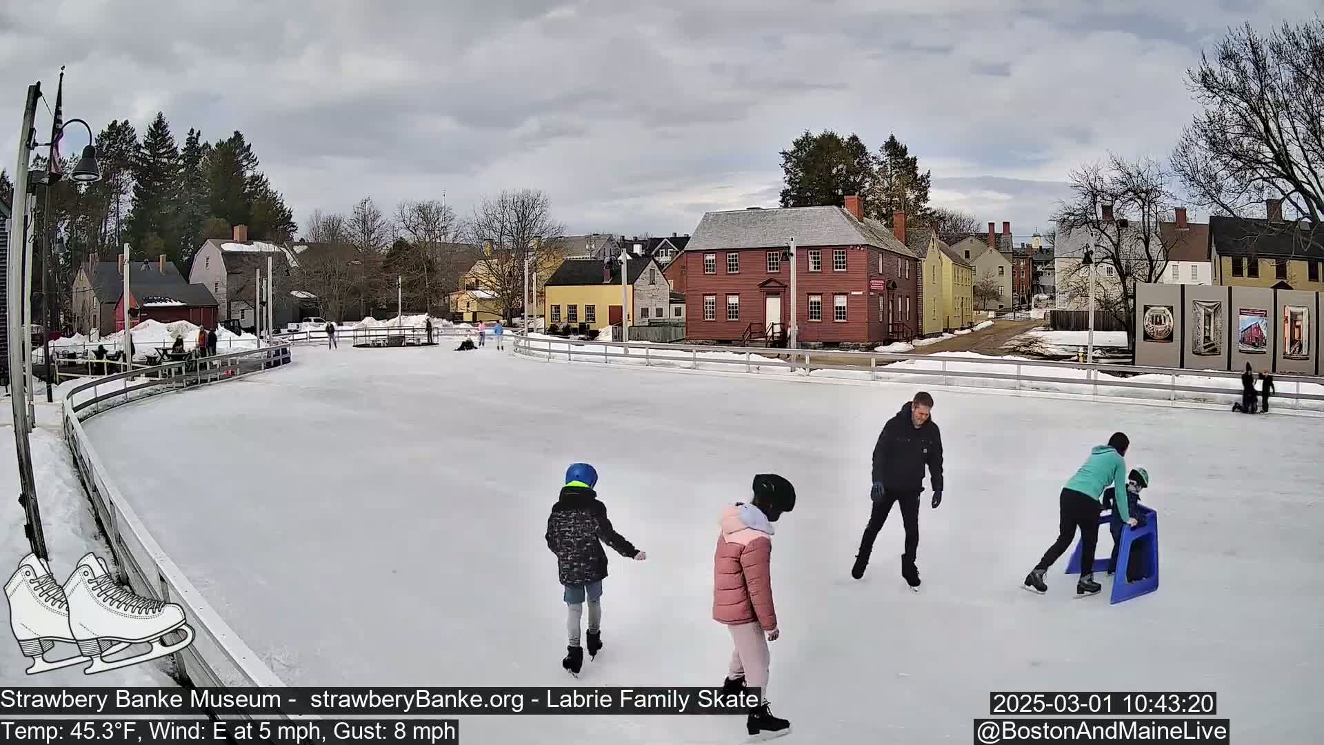Several people ice skate on an outdoor rink in front of historic buildings under a cloudy sky.