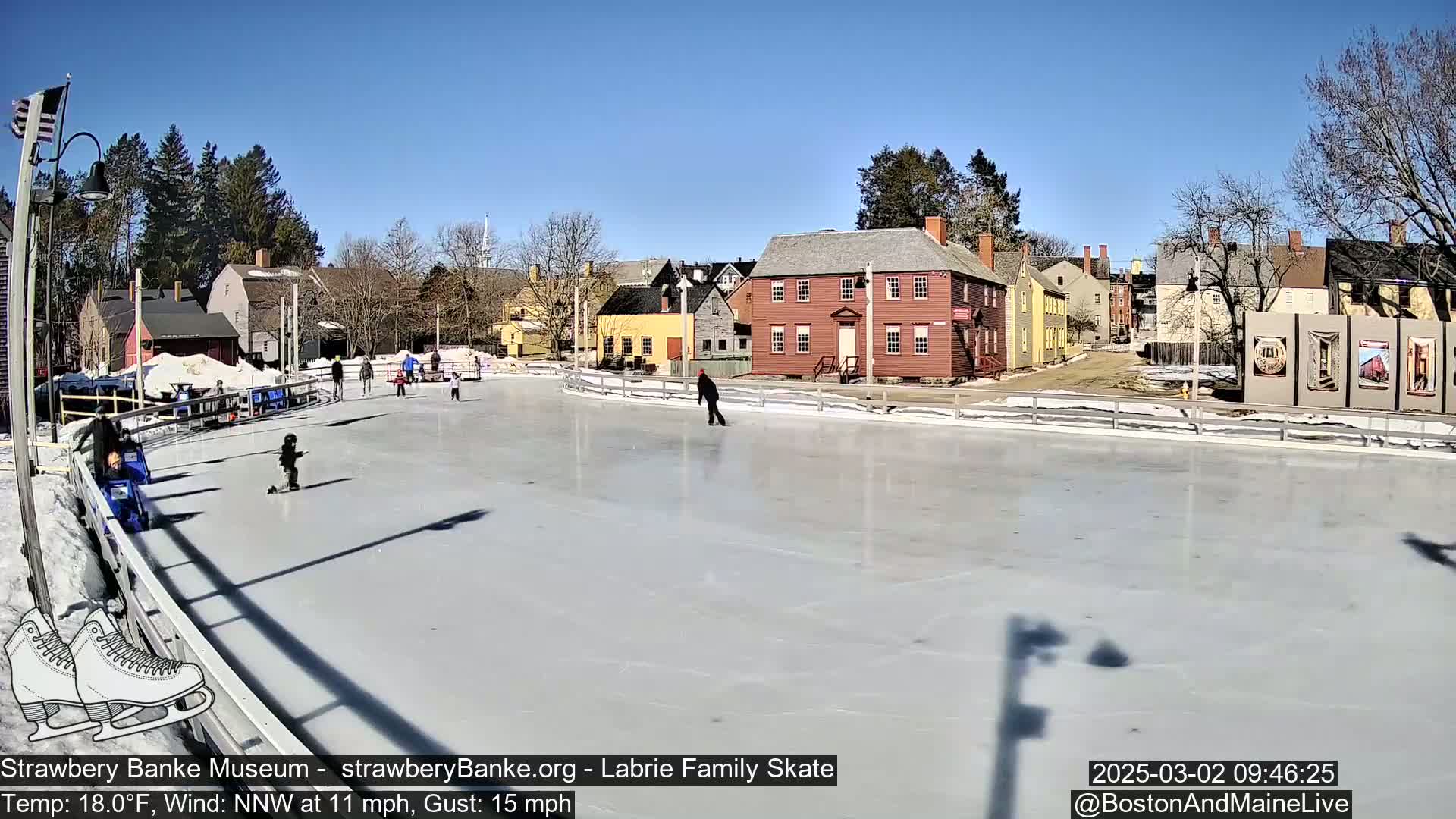 A sunny outdoor ice skating rink, surrounded by historic-looking buildings, is populated by several people ice skating.