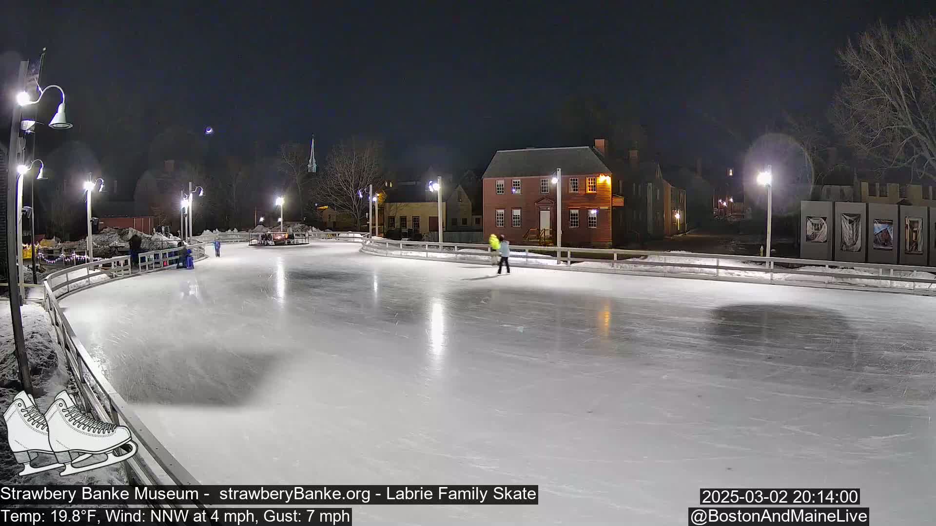 An outdoor ice skating rink, lit by lampposts at night, with a few people ice skating and historical buildings in the background under a clear, dark sky.