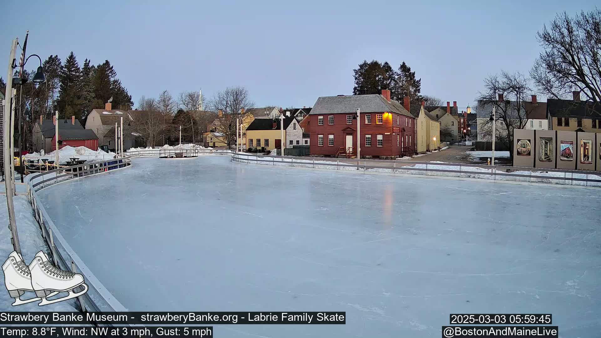 A frozen outdoor skating rink sits in front of a row of historic-looking buildings under a twilight sky.