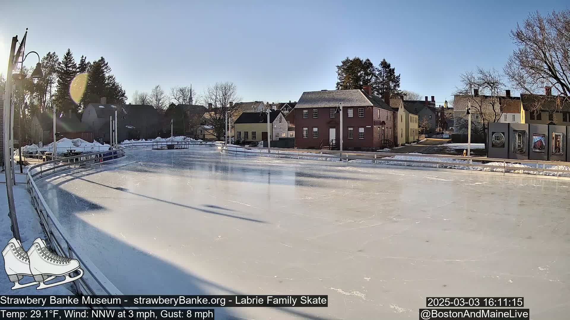 A frozen outdoor skating rink is situated near historical-looking buildings under a sunny sky.