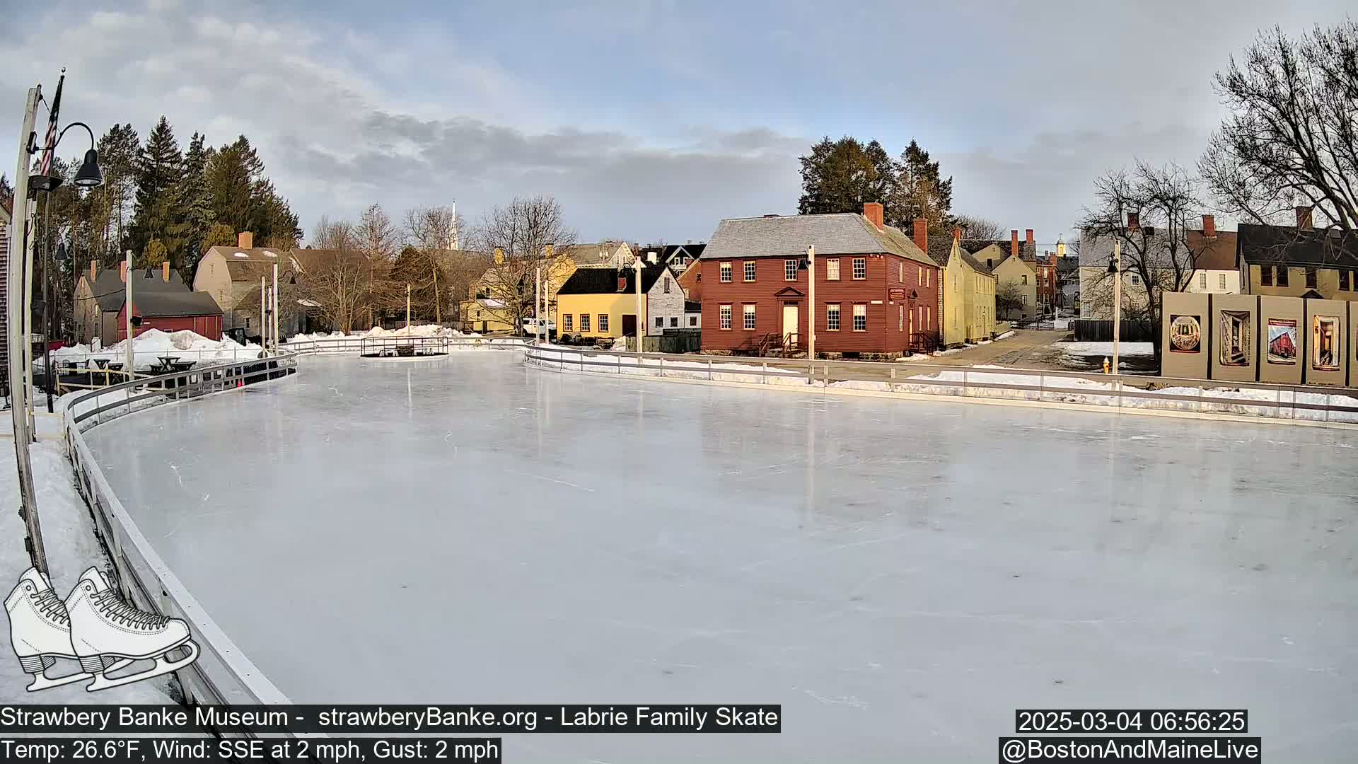 An outdoor ice skating rink sits in front of a row of historic-looking buildings on a partly cloudy day with a light south-southeast wind.