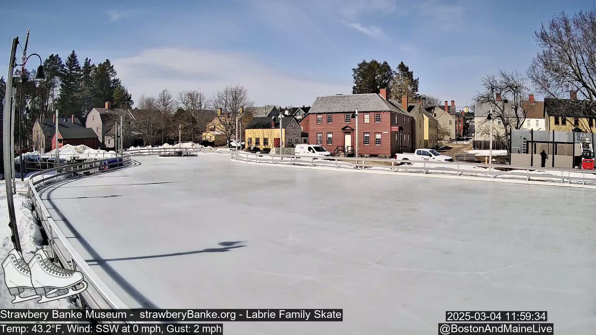An outdoor ice skating rink, surrounded by historic-looking buildings on a sunny day with a light south-southwesterly wind.