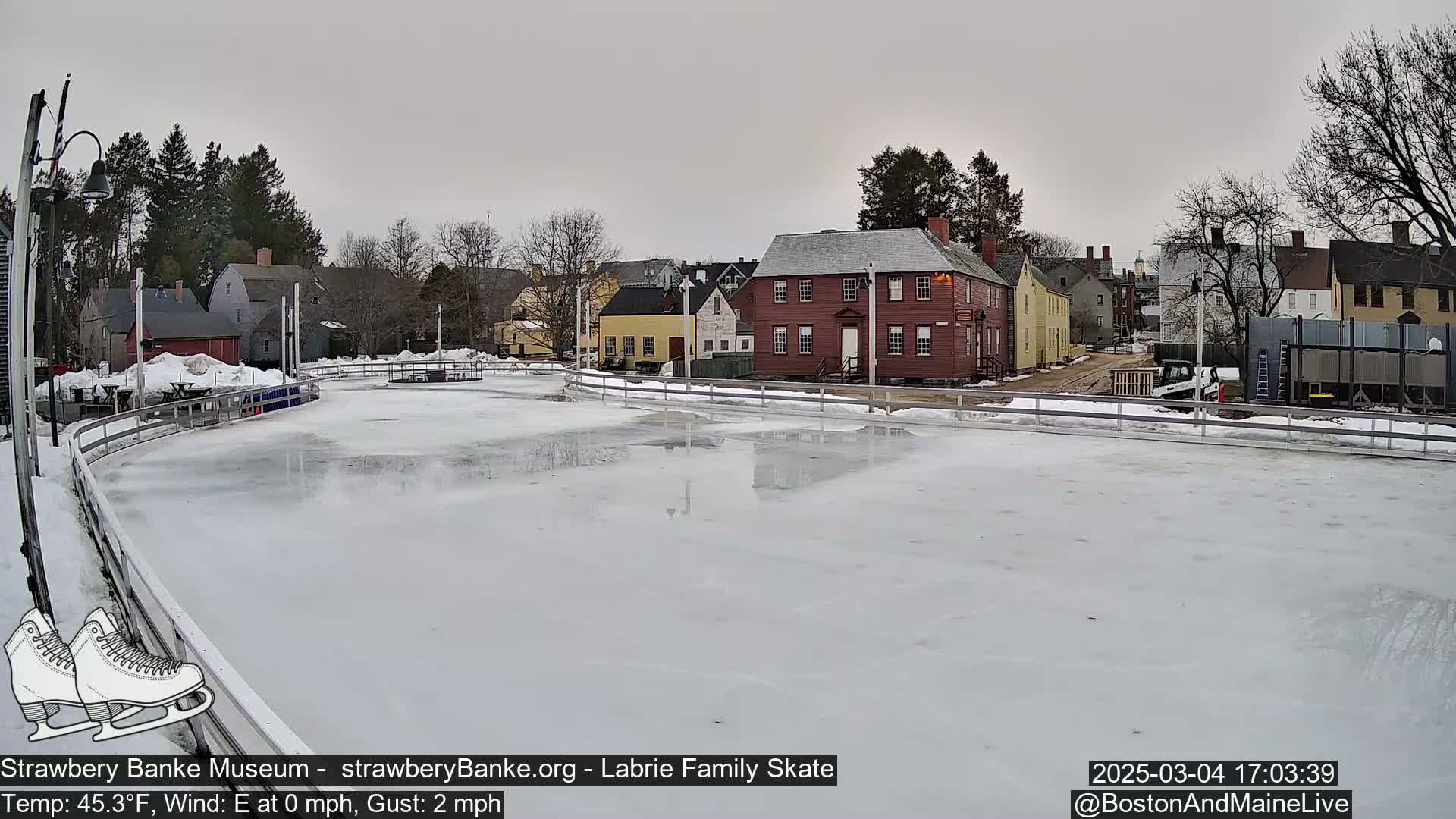 An outdoor ice skating rink, mostly empty, sits in front of a row of historic buildings on an overcast day.