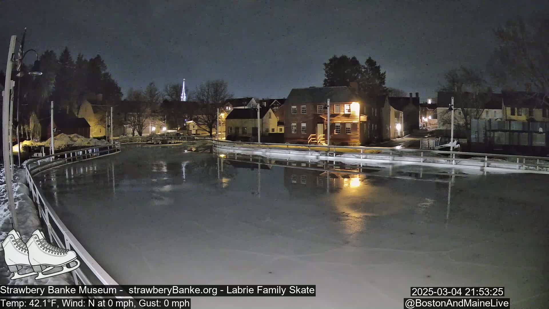 An outdoor ice skating rink at night, illuminated by nearby buildings, reflects the lights onto its calm, dark surface under a clear, dark sky.