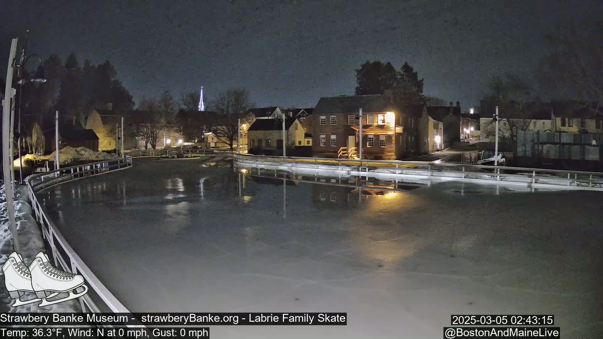 An outdoor ice skating rink at night, illuminated by nearby buildings, with calm weather conditions.