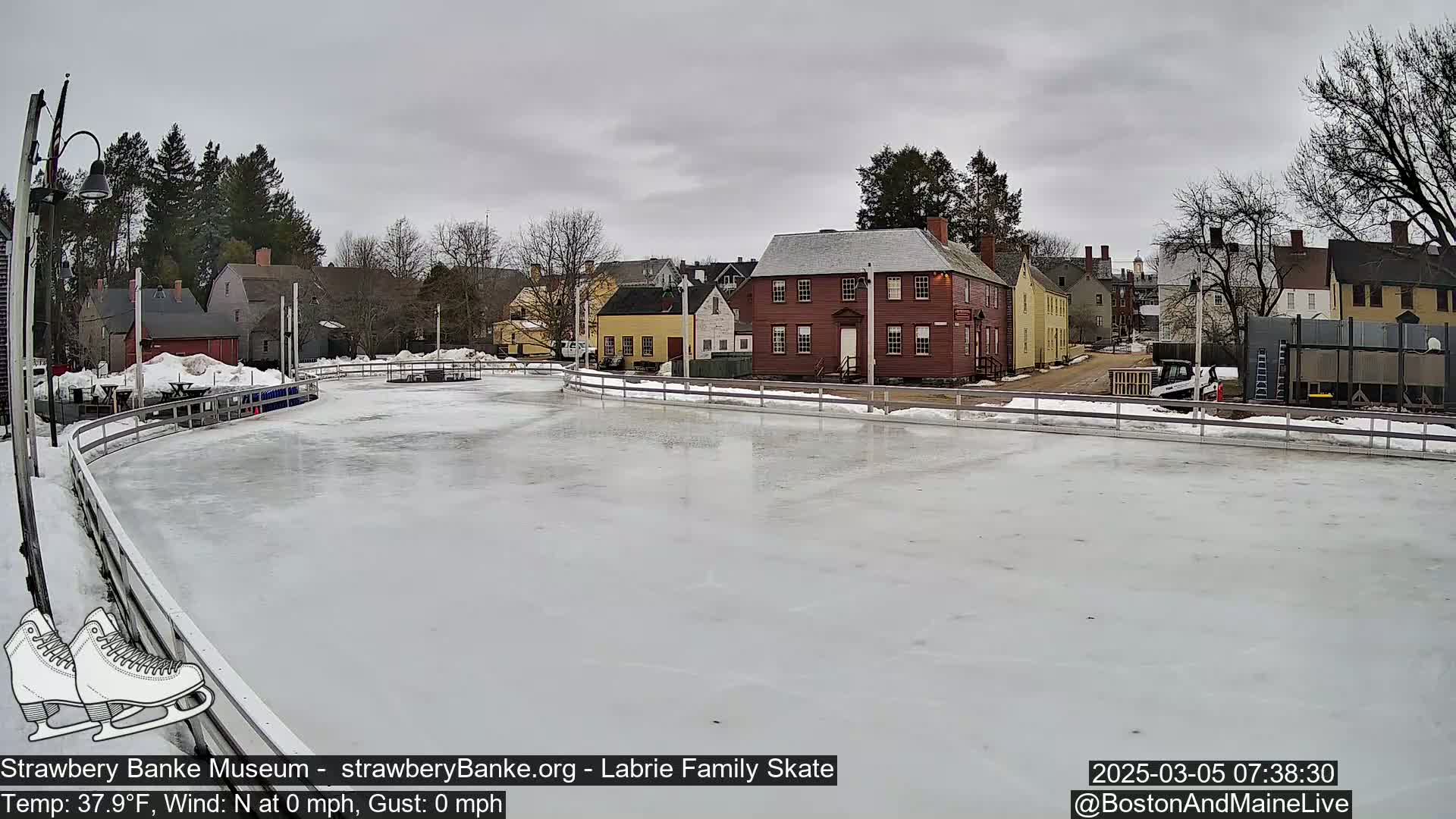 An outdoor ice rink, surrounded by historic buildings under an overcast sky.