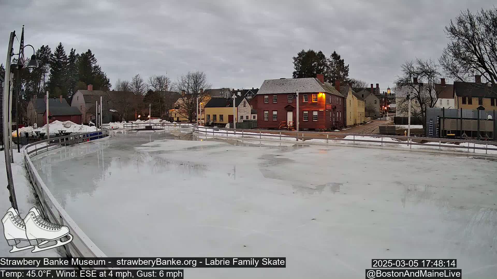 An outdoor ice skating rink, partially melted, sits before a row of historic-looking buildings under an overcast sky.