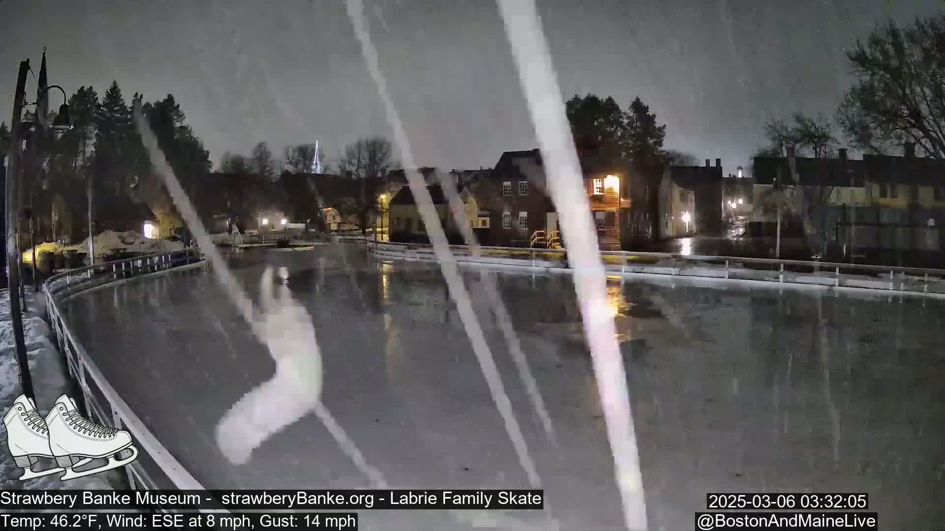 A nighttime view of a rain-slicked outdoor ice skating rink near buildings under a heavy snowfall.