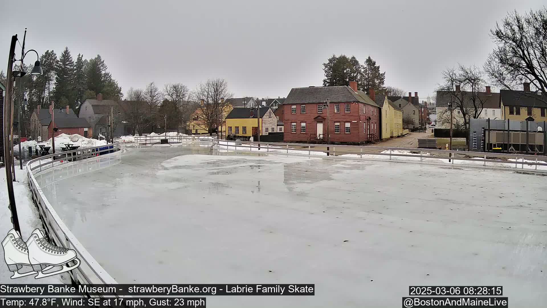 An outdoor ice skating rink, partially covered in snow, sits in front of a row of historic buildings under an overcast sky.