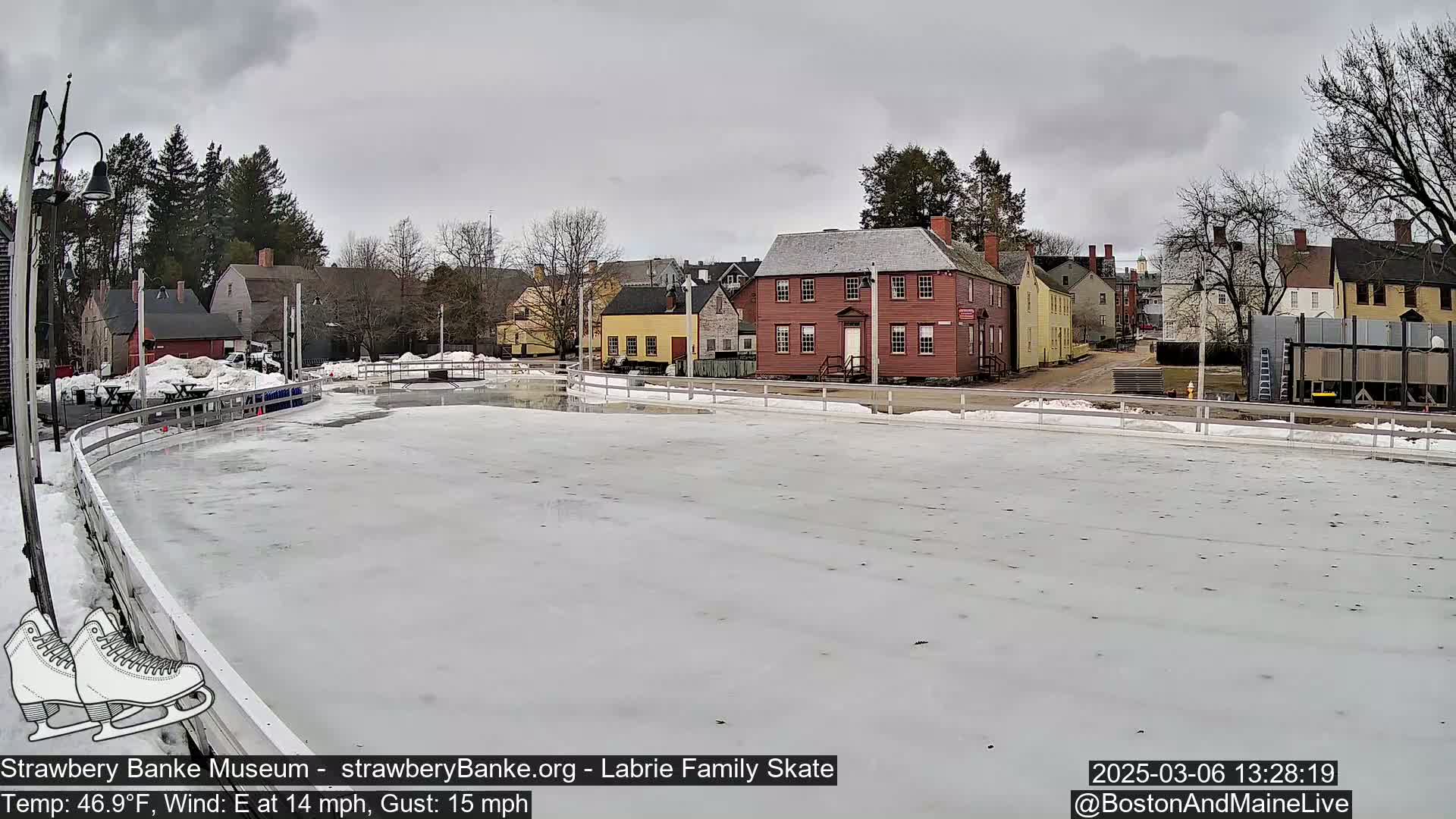 An outdoor ice skating rink is partially covered with snow under an overcast sky, with historic buildings visible in the background.