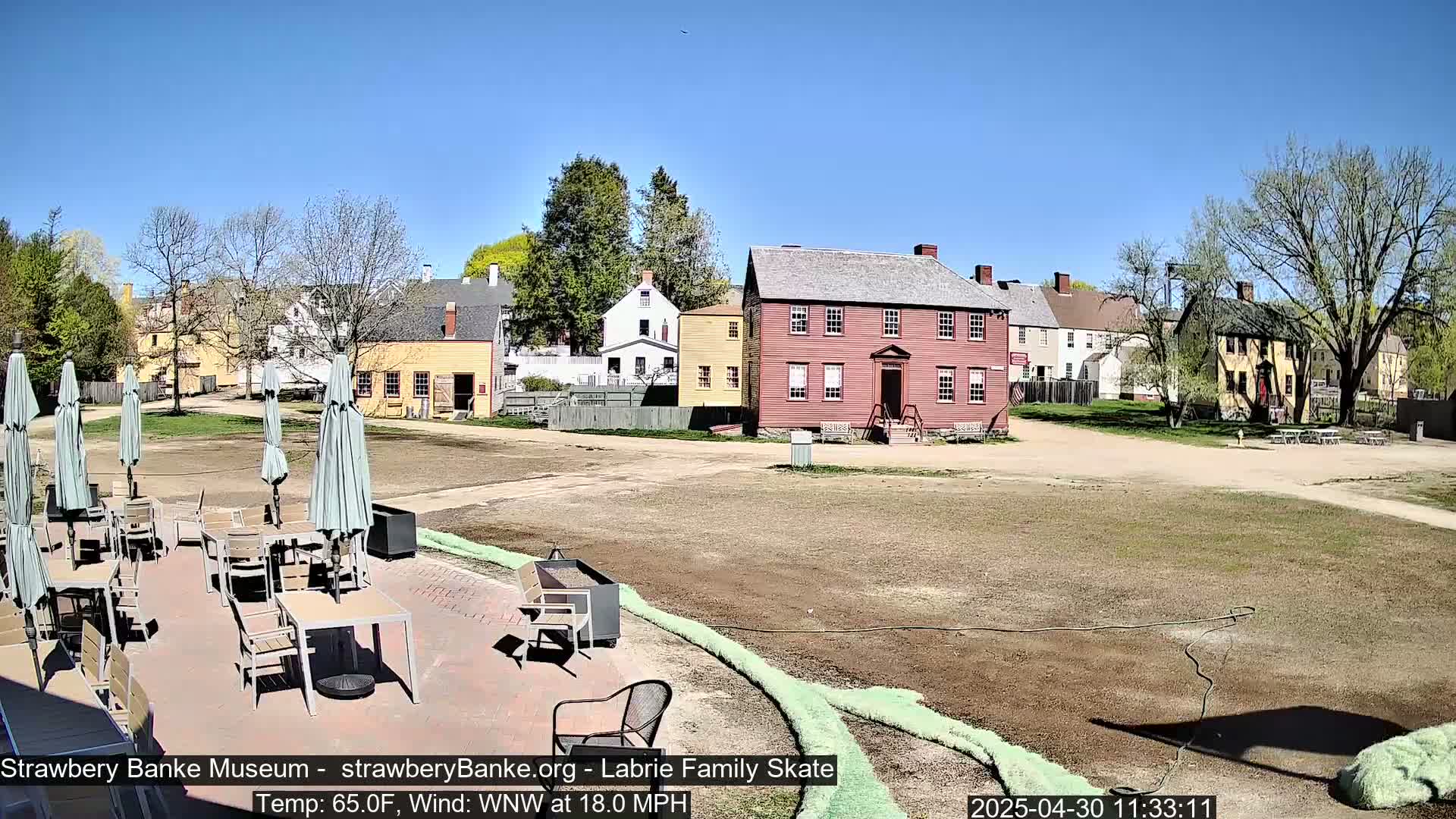 A sunny outdoor scene shows several colonial-era buildings surrounding a grassy courtyard with an outdoor patio furnished with tables and chairs under umbrellas.