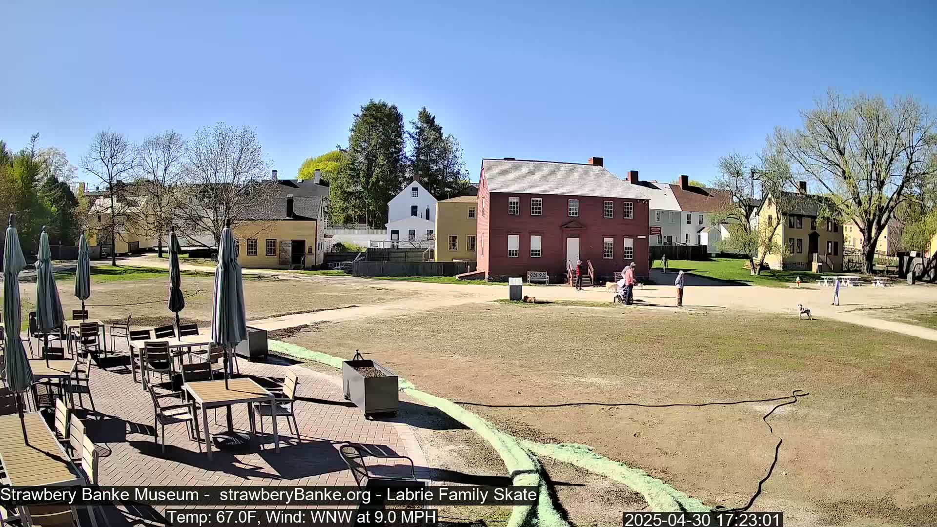 A sunny outdoor scene shows several historic buildings, a grassy area with people and a dog, and a patio with tables and chairs under umbrellas.