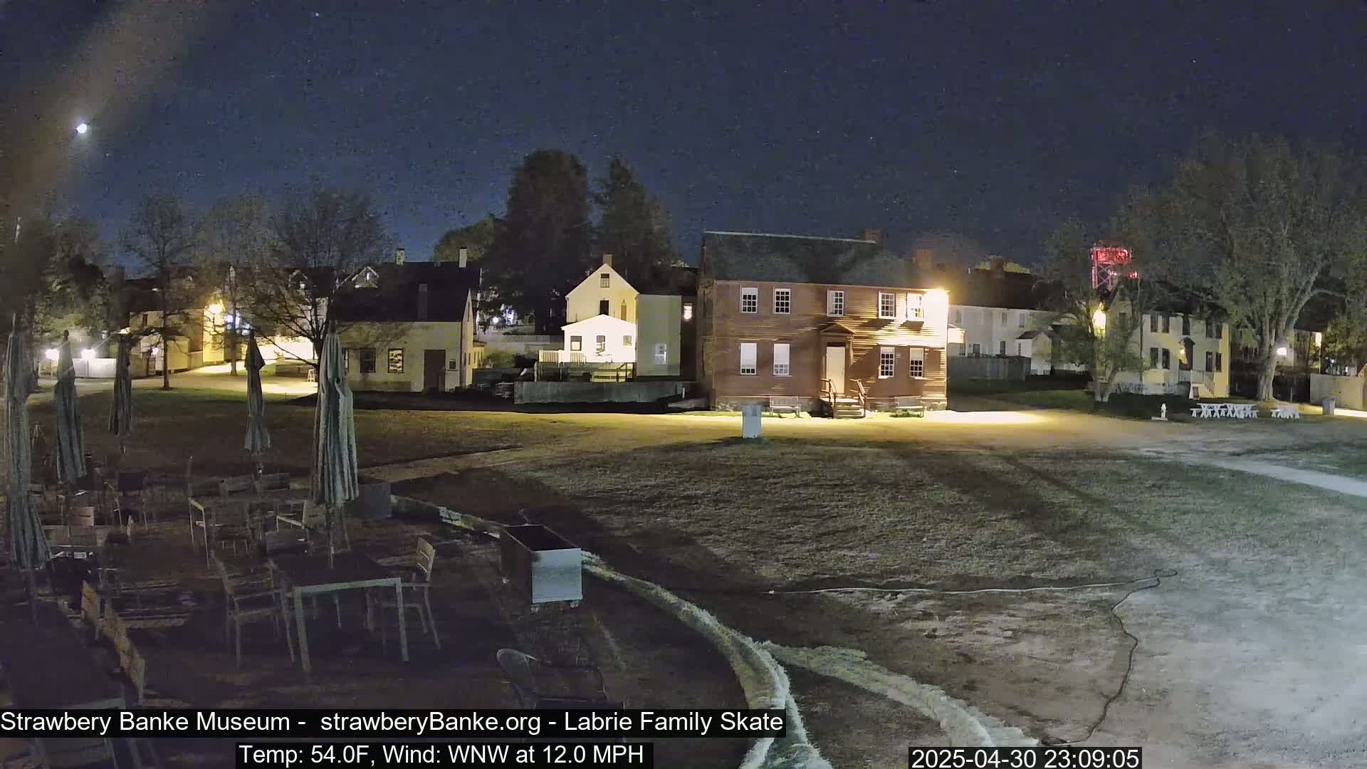 A nighttime view of several colonial-style buildings and a grassy area with outdoor furniture, under a clear, starry sky.