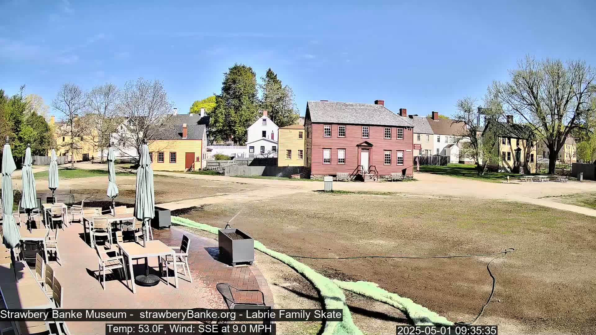 A patio with tables and umbrellas sits in front of a collection of historic-looking buildings on a sunny day with a light wind.