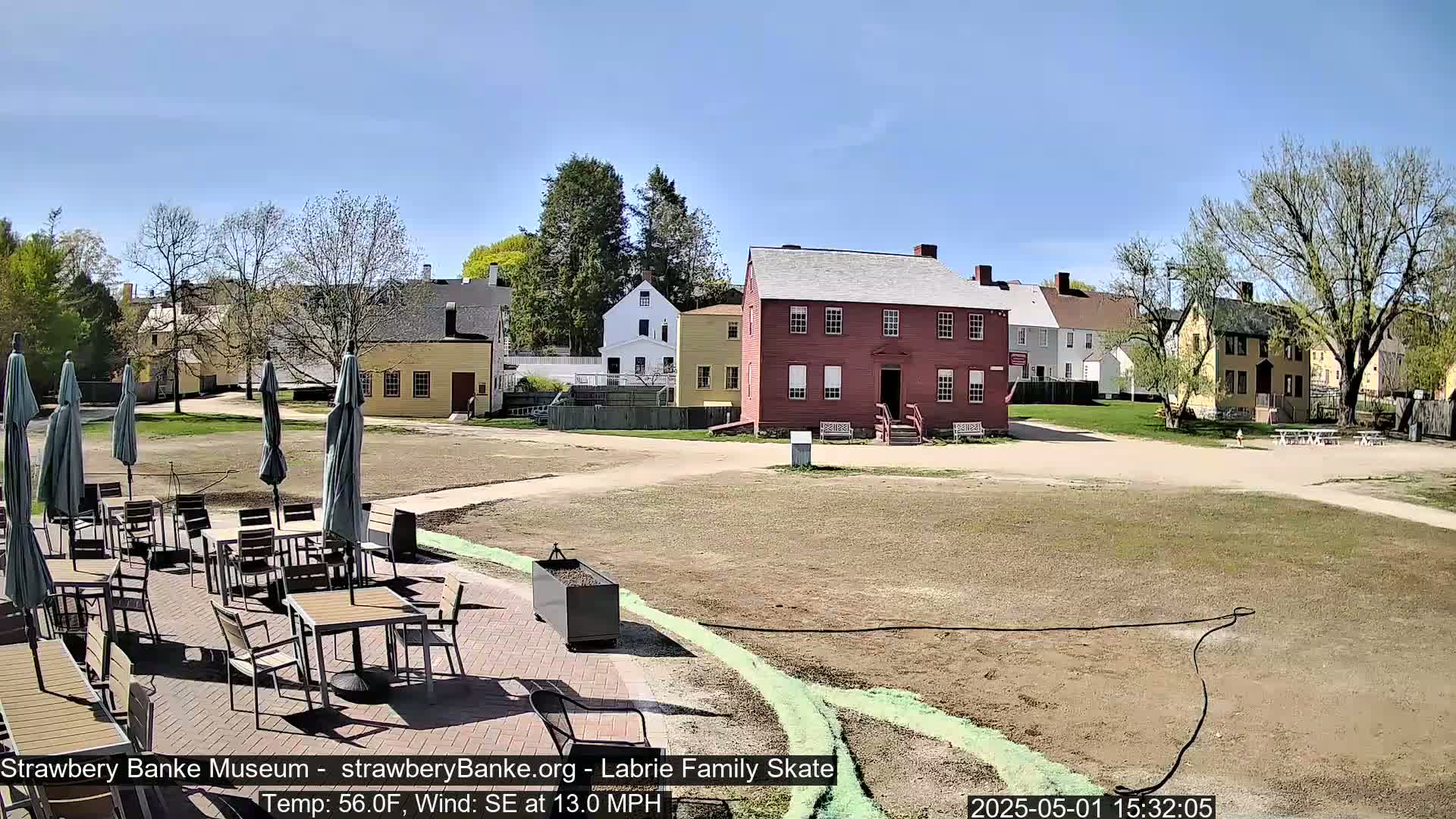 A sunny outdoor scene shows a collection of historic-looking buildings and a patio area with tables and umbrellas.