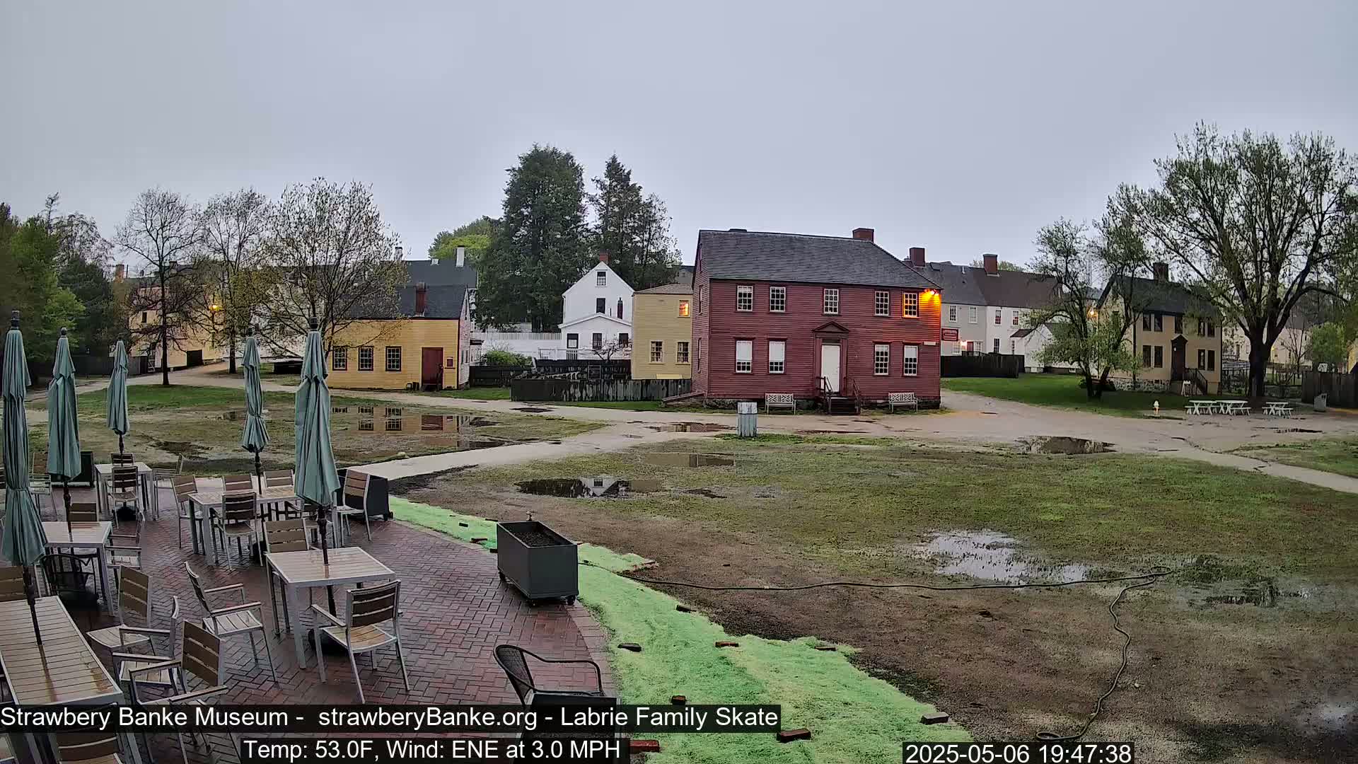 A courtyard with several colonial-style buildings, outdoor seating with umbrellas, and puddles of water under an overcast sky.