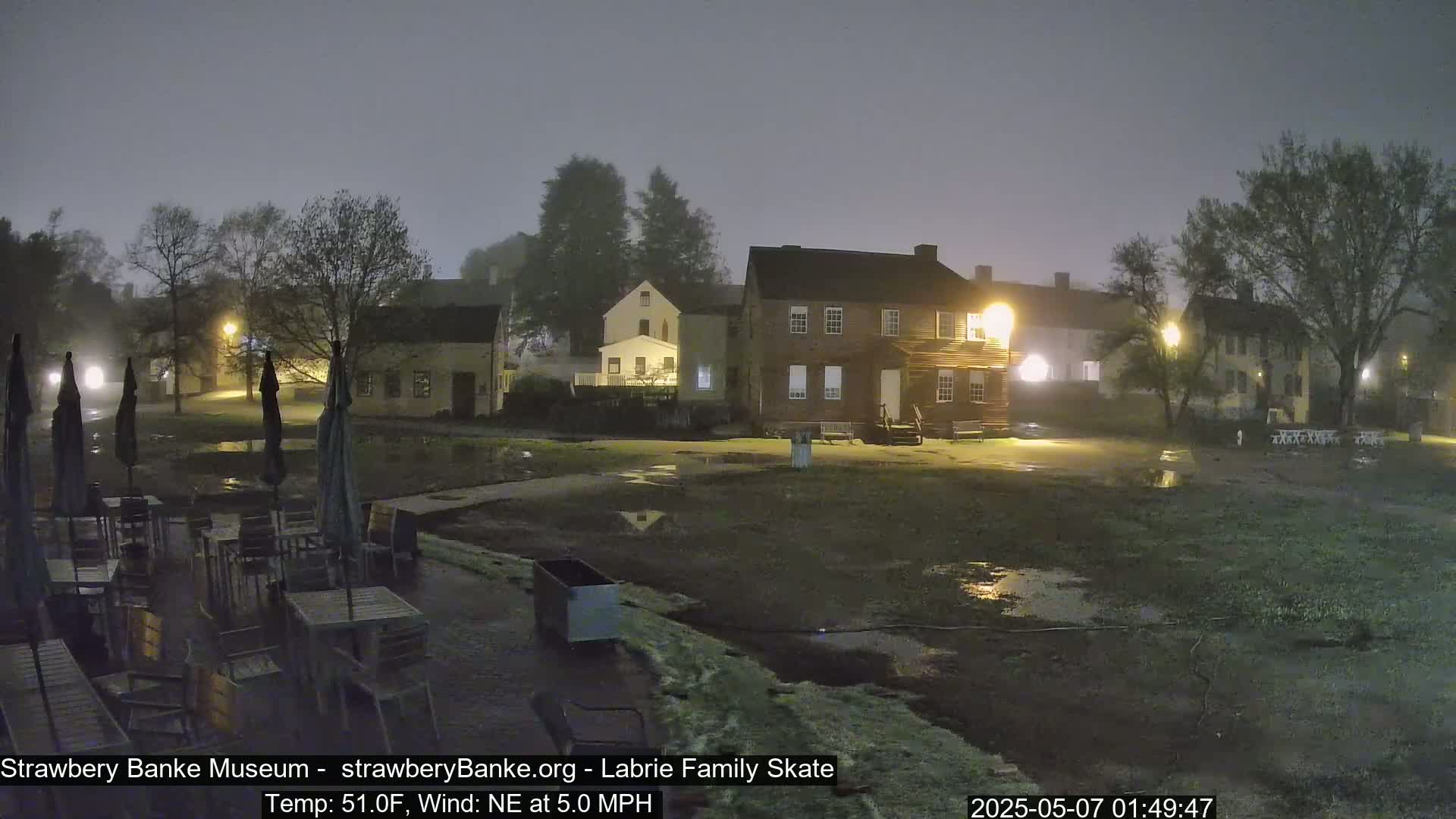 A fog-shrouded nighttime scene shows several historic buildings and an outdoor patio area with tables and umbrellas, all wet from a light rain or mist.