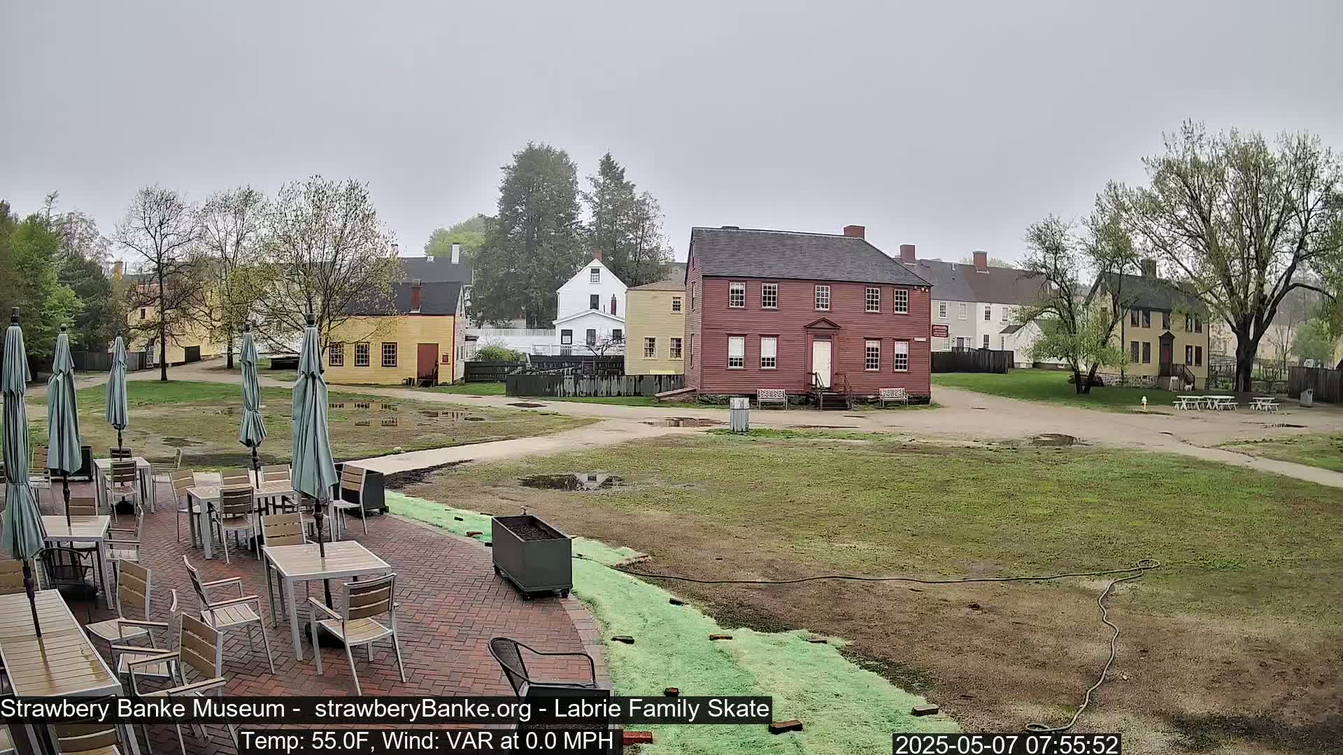 A patio with tables and umbrellas overlooks a grassy area and several historic-looking buildings on an overcast day.