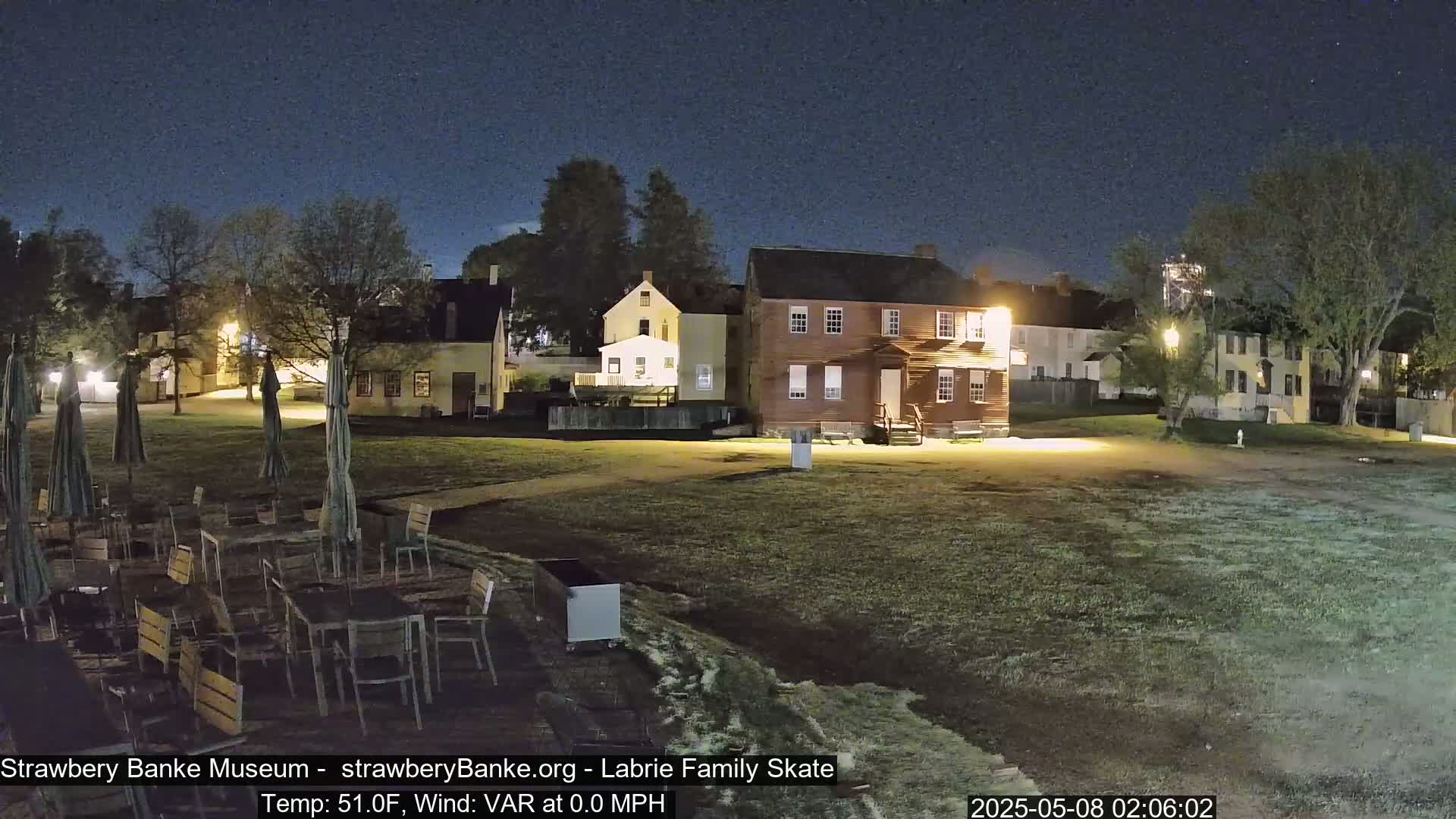 A nighttime view of a grassy area with several historical-looking buildings, outdoor seating with closed umbrellas, and calm weather conditions.