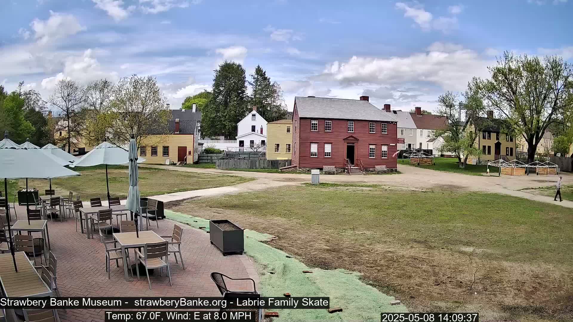 A patio with tables and umbrellas overlooks a grassy area and several historic-looking buildings on a partly cloudy day with an east wind.