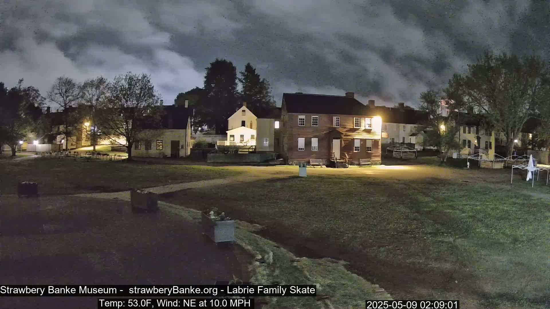 Under a cloudy night sky, several historical-looking buildings are illuminated on a grassy area, with a gentle northeast wind blowing at 10 mph and a temperature of 53°F.