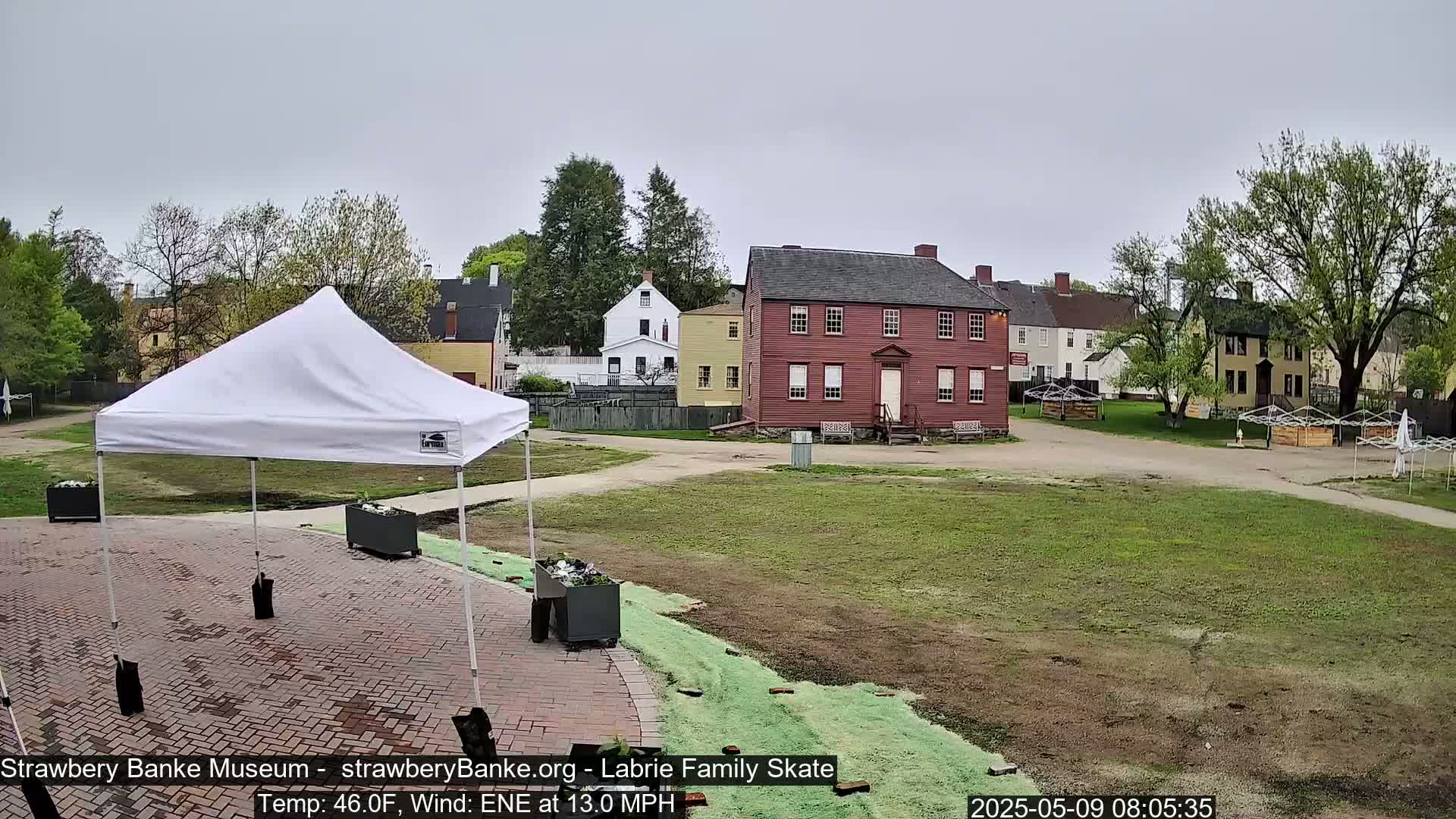 A white canopy tent stands in a brick paved area, overlooking a grassy field with several historical-looking buildings in the background on an overcast day.