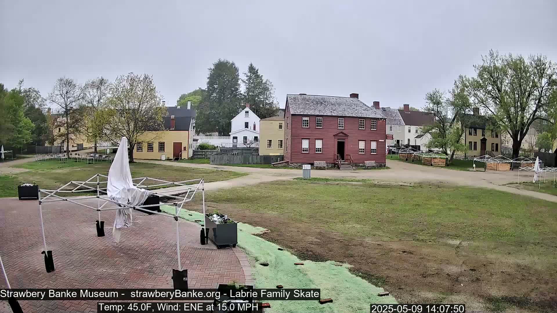 A mostly overcast outdoor scene shows a collection of historic-looking buildings on a grassy area, with several covered tables and a partially covered canopy in the foreground.