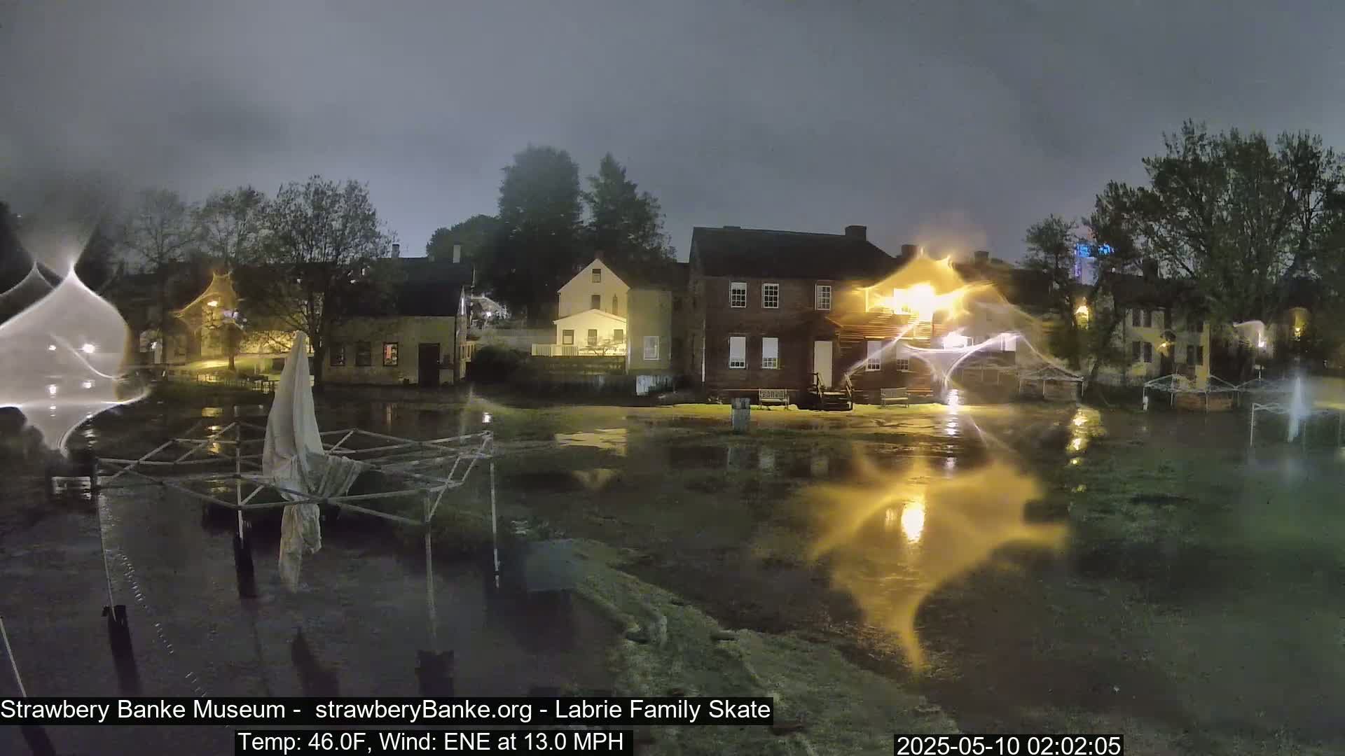 Floodwaters illuminate several colonial-style buildings at night under a dark, overcast sky with a 13 mph wind.
