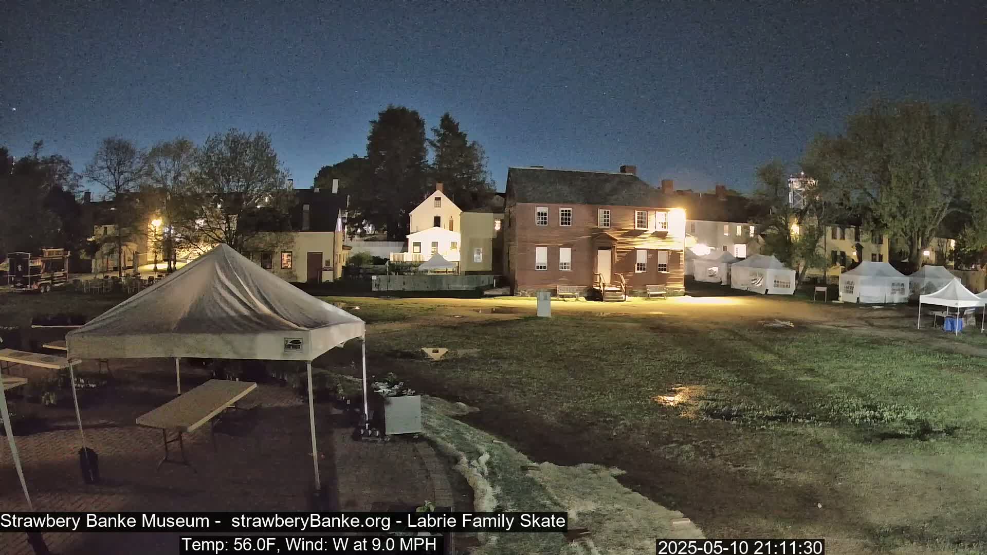 Under a night sky, several tents and historic-looking buildings are illuminated in a damp, grassy area with puddles.
