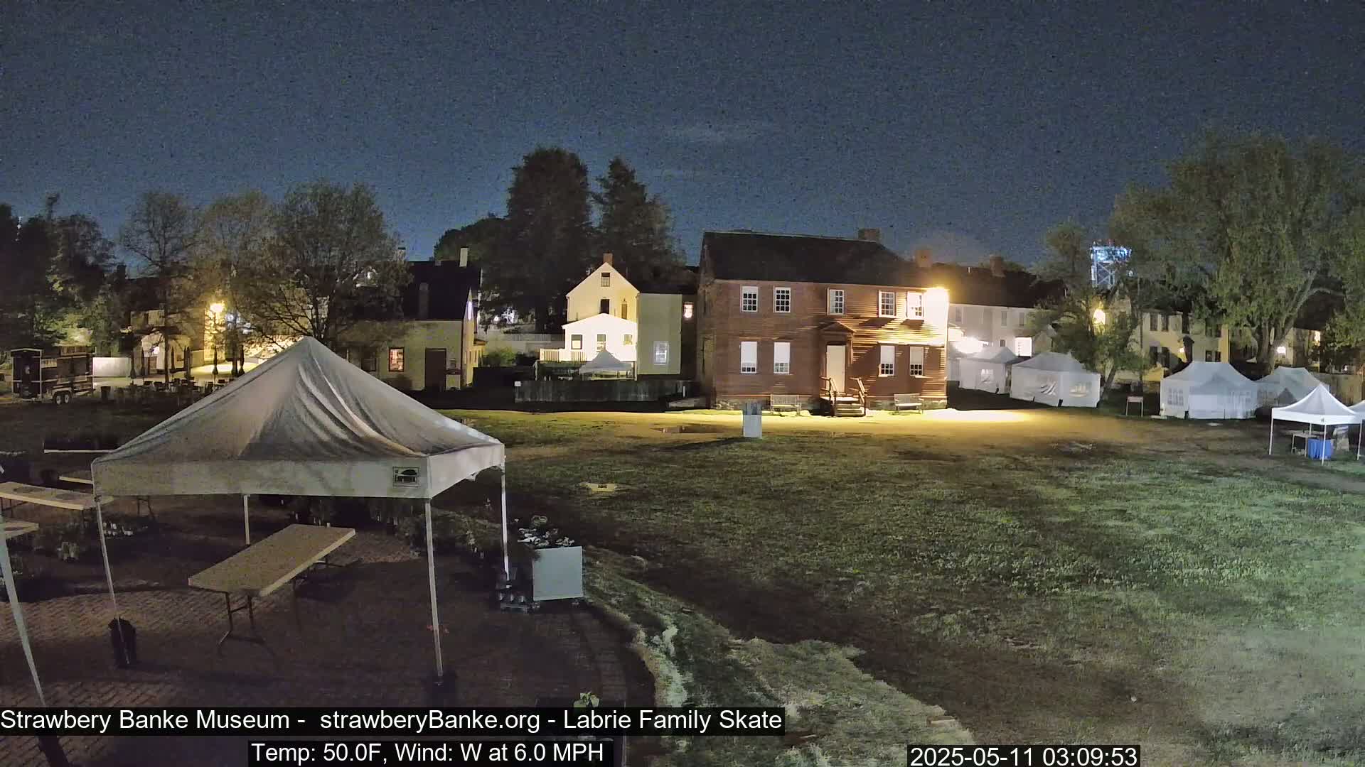 At night, under a clear sky, a grassy area is illuminated, showing several tents, some buildings, and a white canopy tent in the foreground.