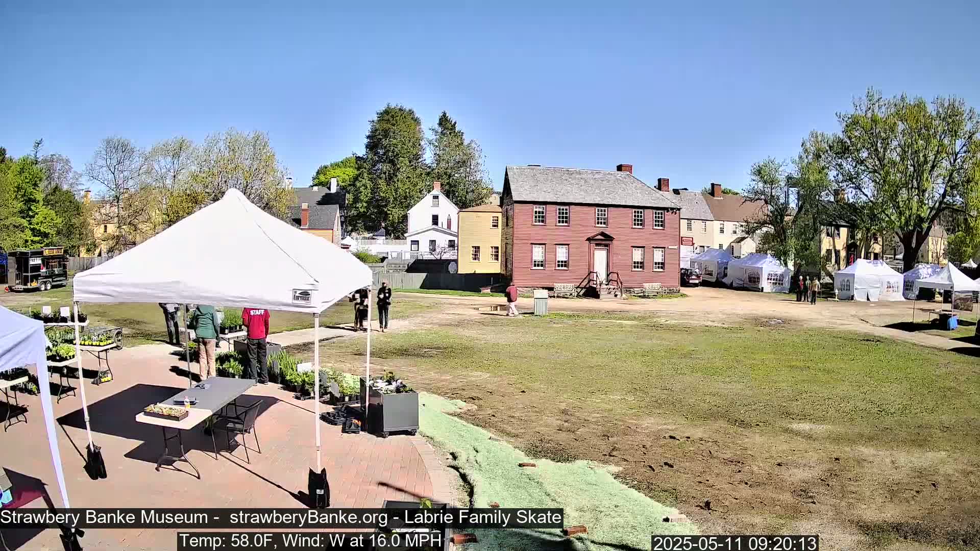 A sunny outdoor scene shows several white tents set up on a grassy area near historical-looking buildings, with a few people milling about.