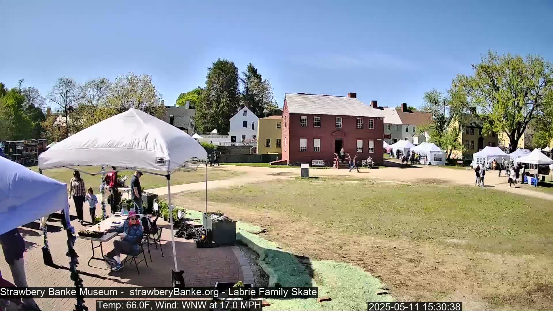 A sunny outdoor scene shows a grassy area with several white tents, a red brick building, and other historical-looking structures, with people walking around.