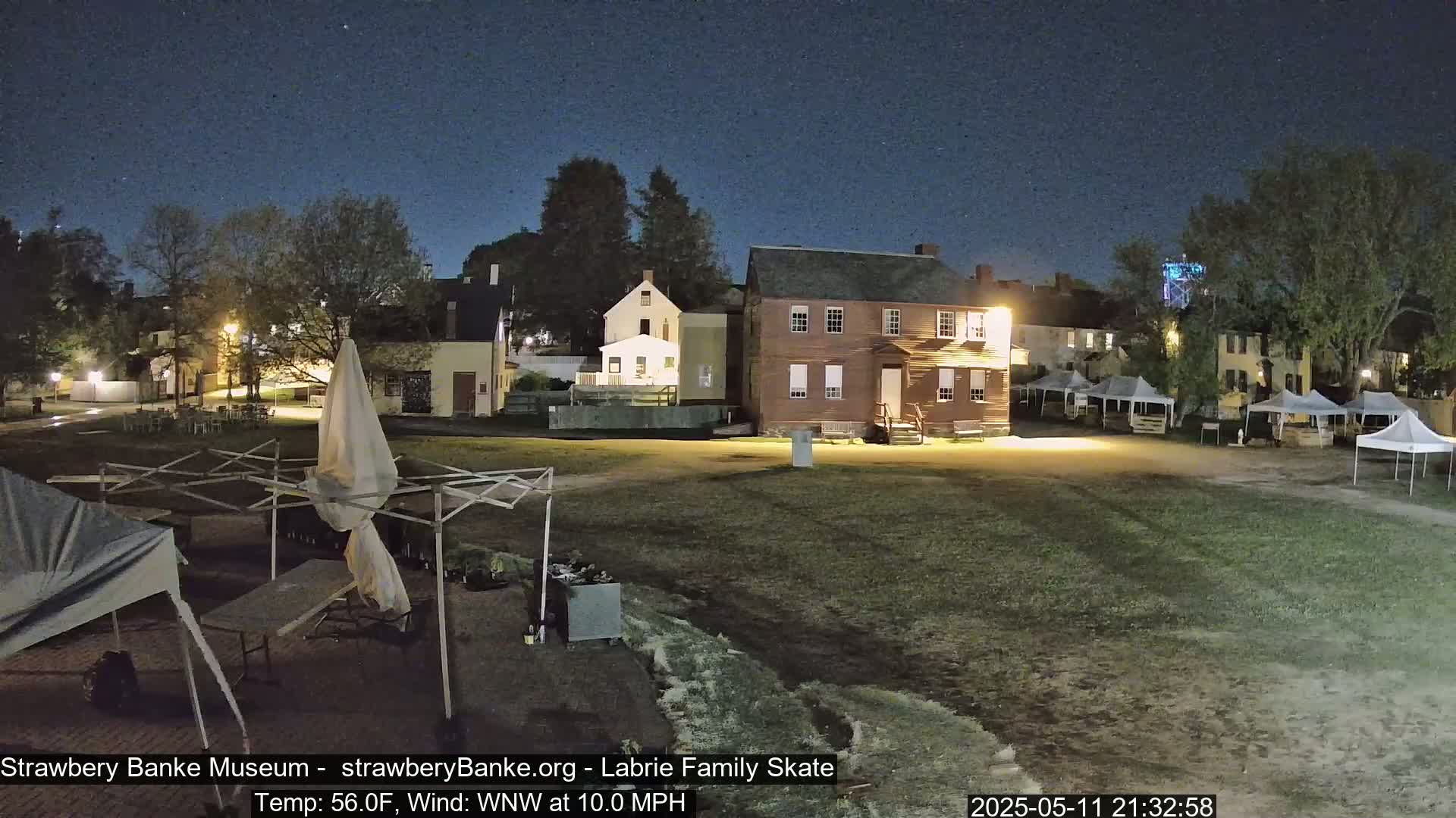 A nighttime view of a grassy area with several historical-looking buildings, some tents, and trees under a clear, dark sky.