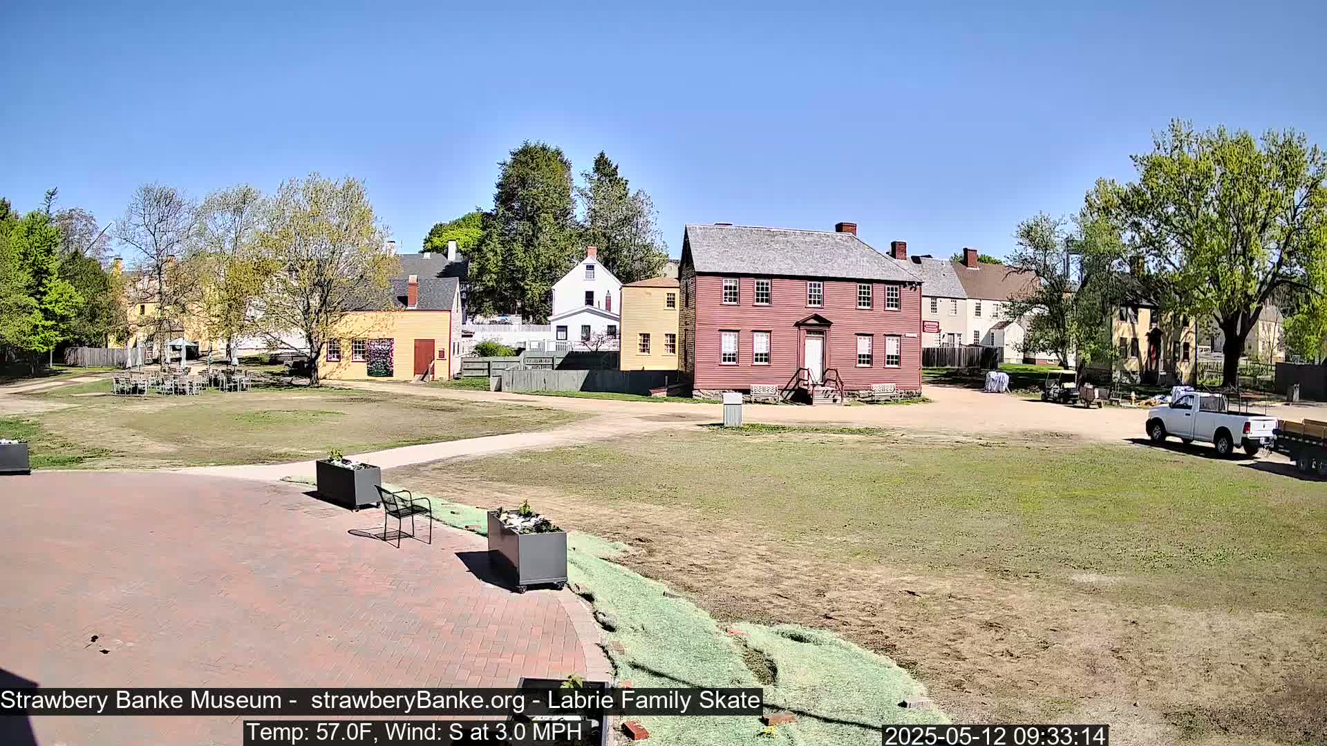 A sunny outdoor scene shows several historic-looking buildings, a grassy area with some seating, and a pickup truck, under a clear blue sky.